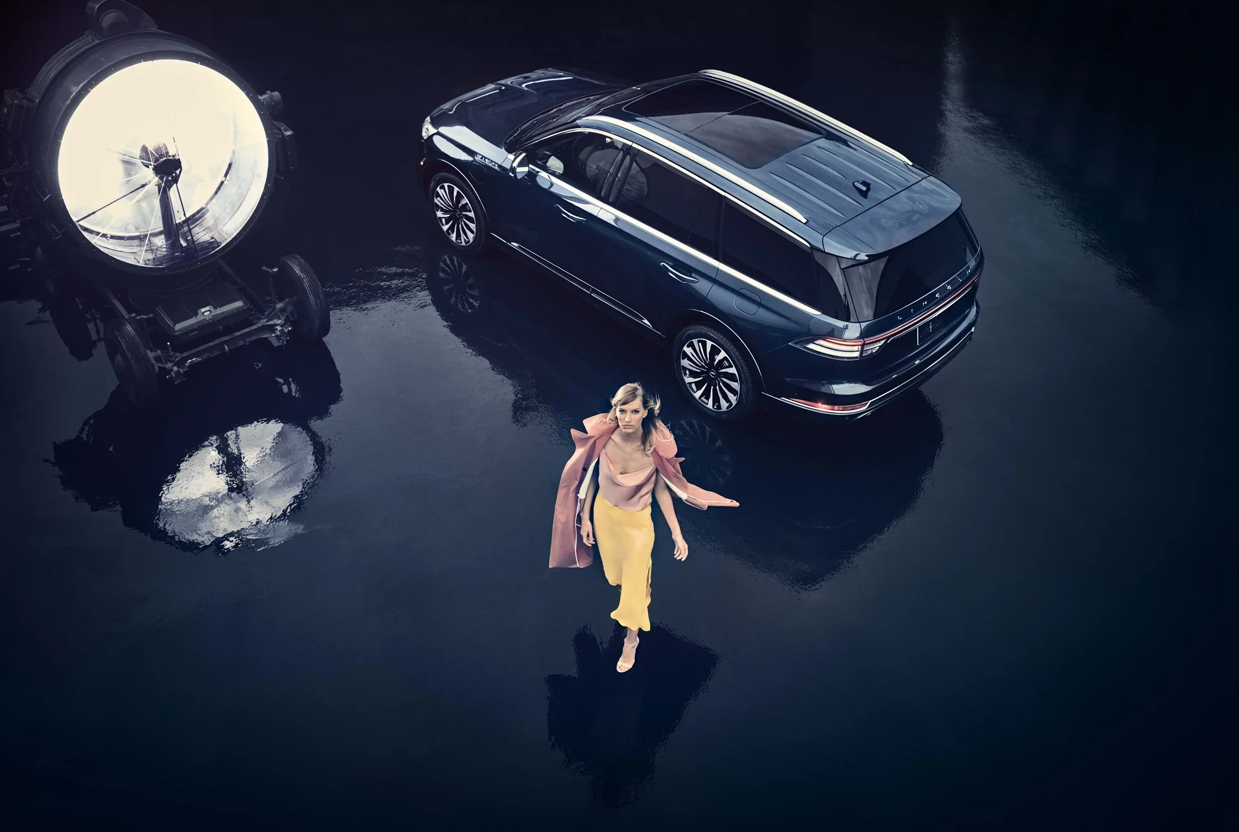 A woman in a pink jacket and yellow pants walking on a wet surface near a black luxury station wagon car, with a large studio light to the left reflecting on the ground. Lincoln Continental from Lincoln Motor Company.  