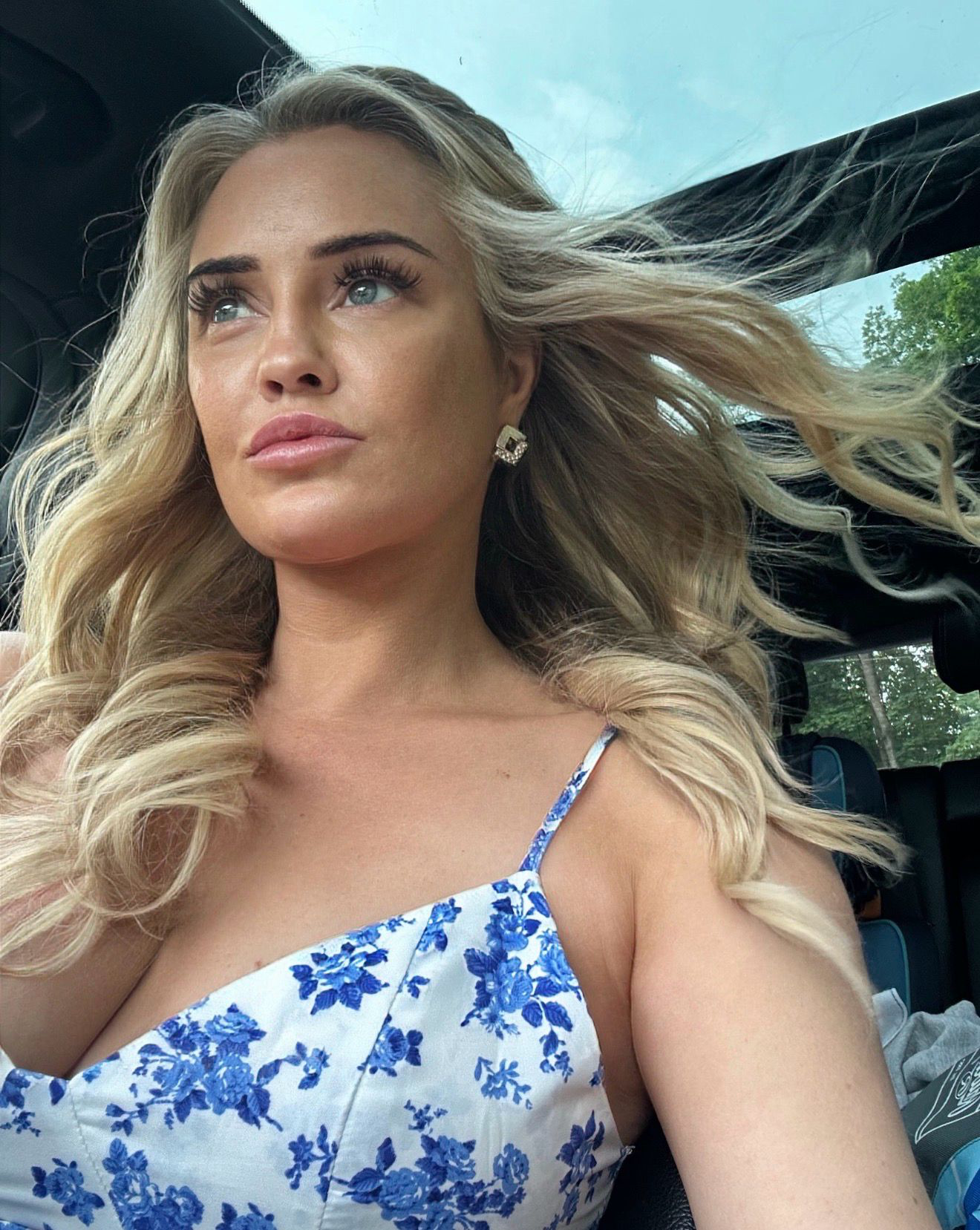 Close-up of a young woman with long wavy blonde hair, blue eyes, full lips, and wearing a white top, against a plain light background.