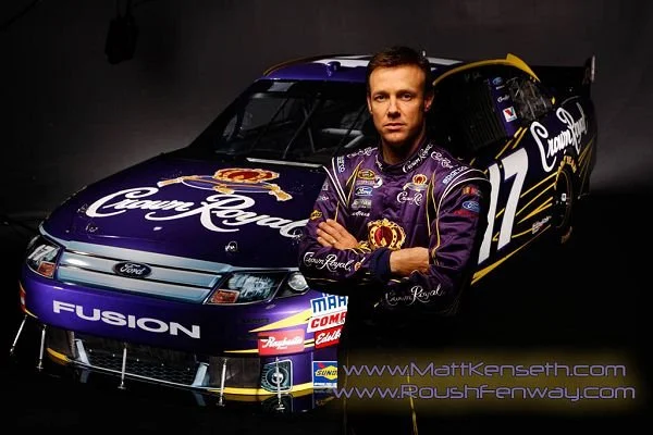 A man in a purple racing suit standing with arms crossed in front of a purple NASCAR race car with the Crown Royal logo, number 17, and sponsor decals.