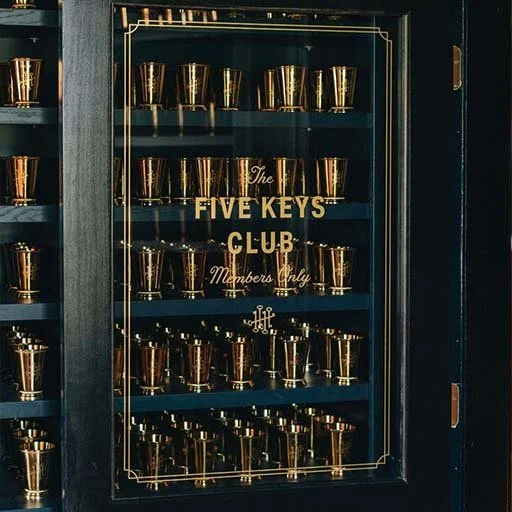 A dark cabinet containing multiple gold-colored cups organized on shelves behind a glass door, with gold labeling reading 'The Five Keys Club Members Only' in the center.