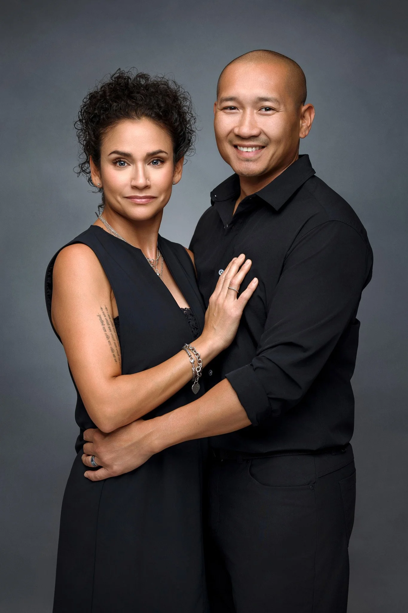A smiling man and woman pose together in front of a gray backdrop, dressed in black clothing.