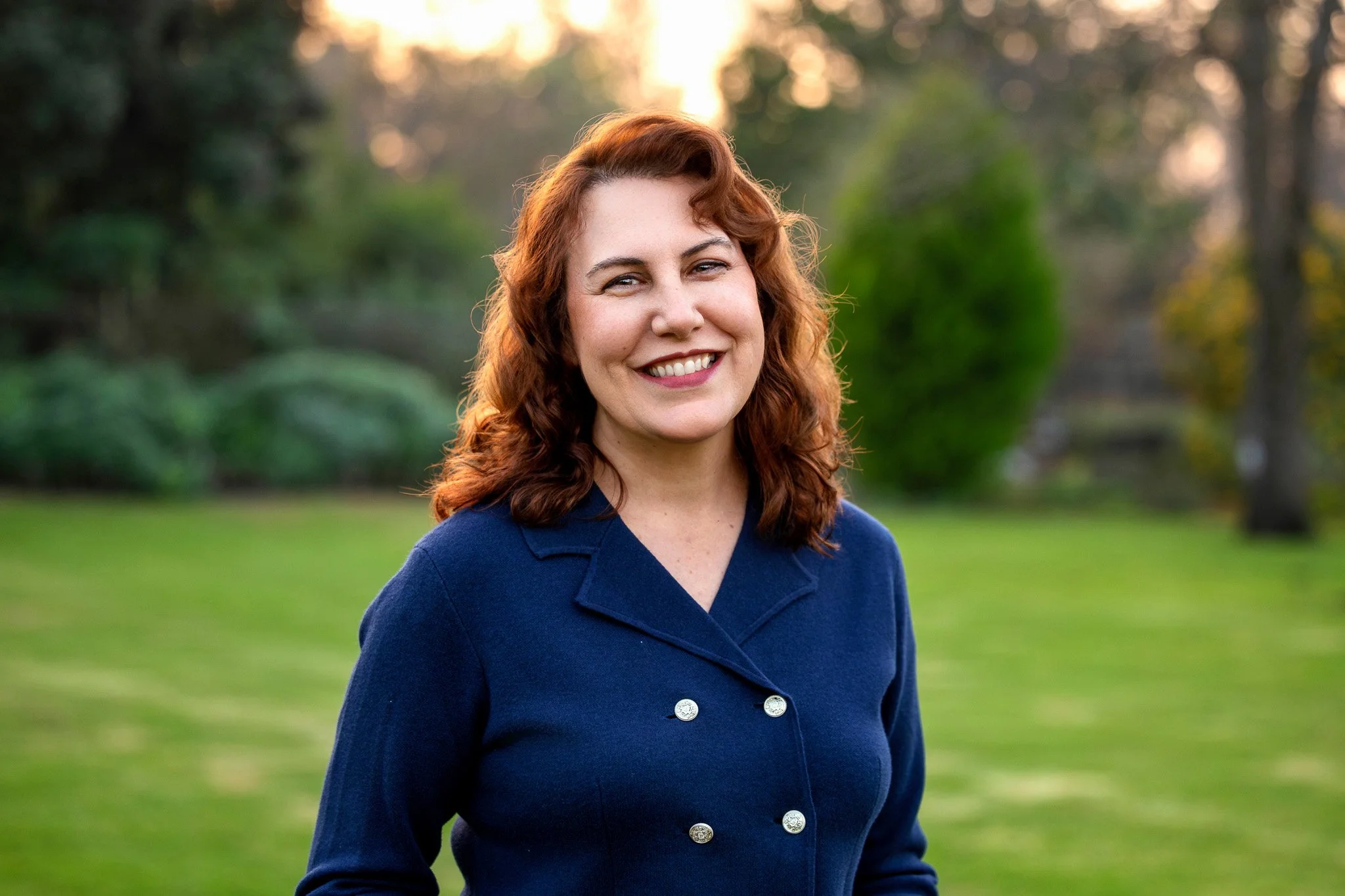 A woman with curly red hair, smiling, wearing a navy blue coat with silver buttons, standing outdoors in a park-like setting during sunset.