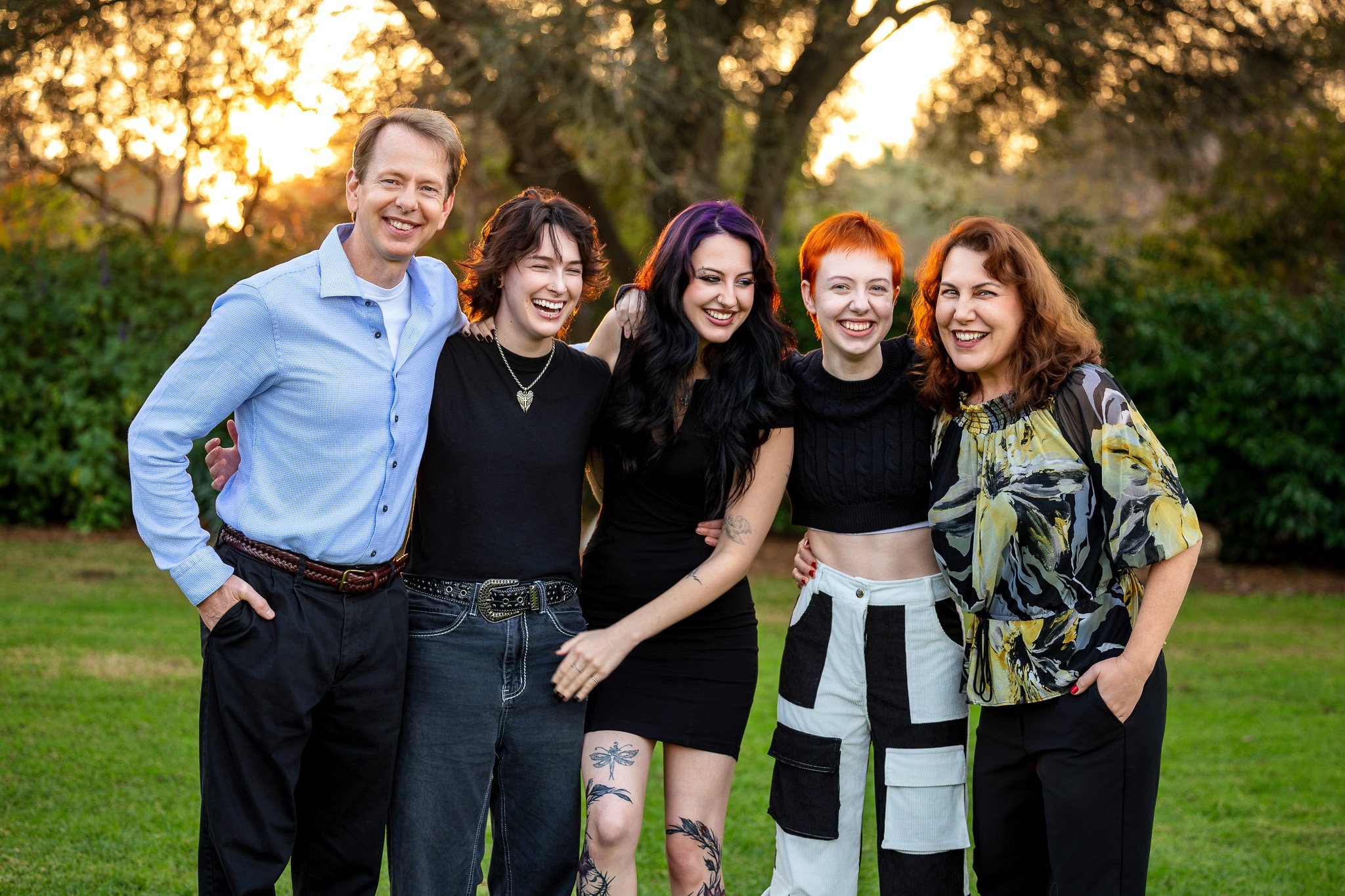 A group of five diverse young adults posing together outdoors during sunset, smiling and embracing each other.