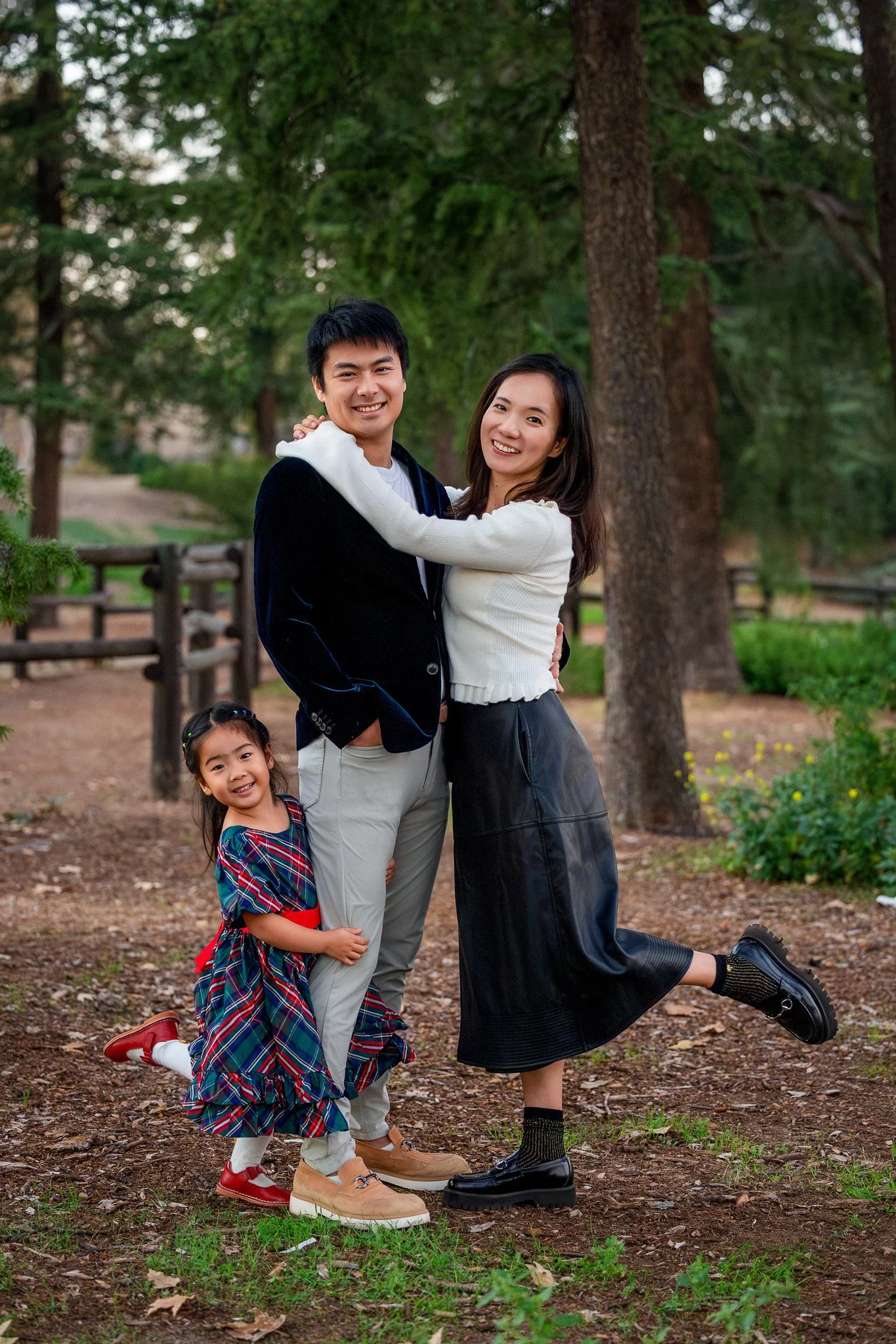 A happy family of three, a young man, a young woman, and a young girl, standing outdoors on a dirt path in a park with tall trees. The girl is wearing a colorful plaid dress, and the adults are smiling and embracing each other.
