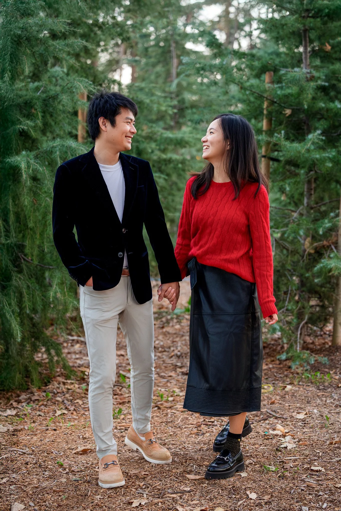 A young couple holding hands and smiling at each other while walking through a forest with green trees and leaves on the ground.