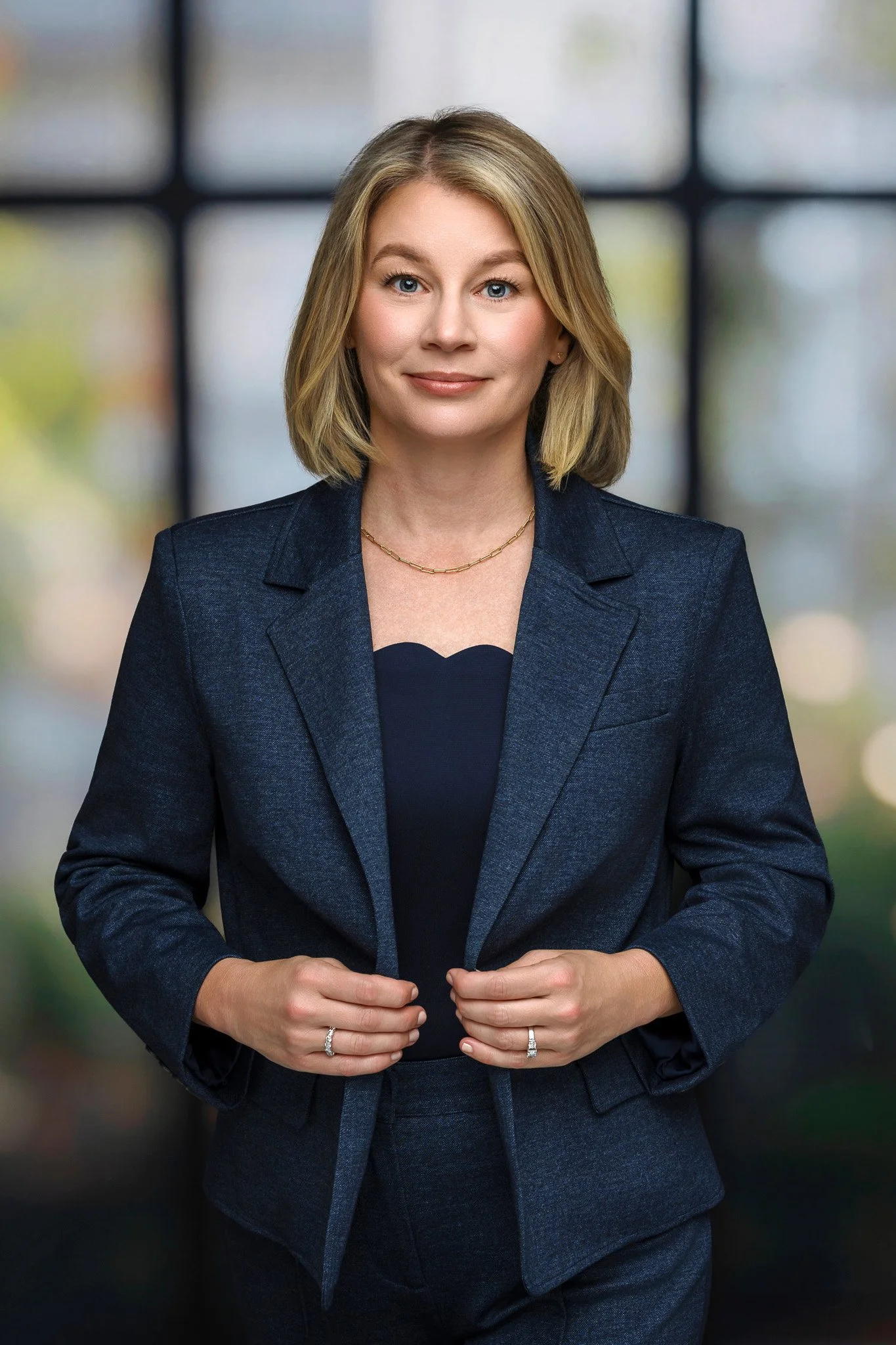 A professional woman with shoulder-length blonde hair wearing a dark blue blazer and matching skirt, standing indoors in front of a blurred window background.
