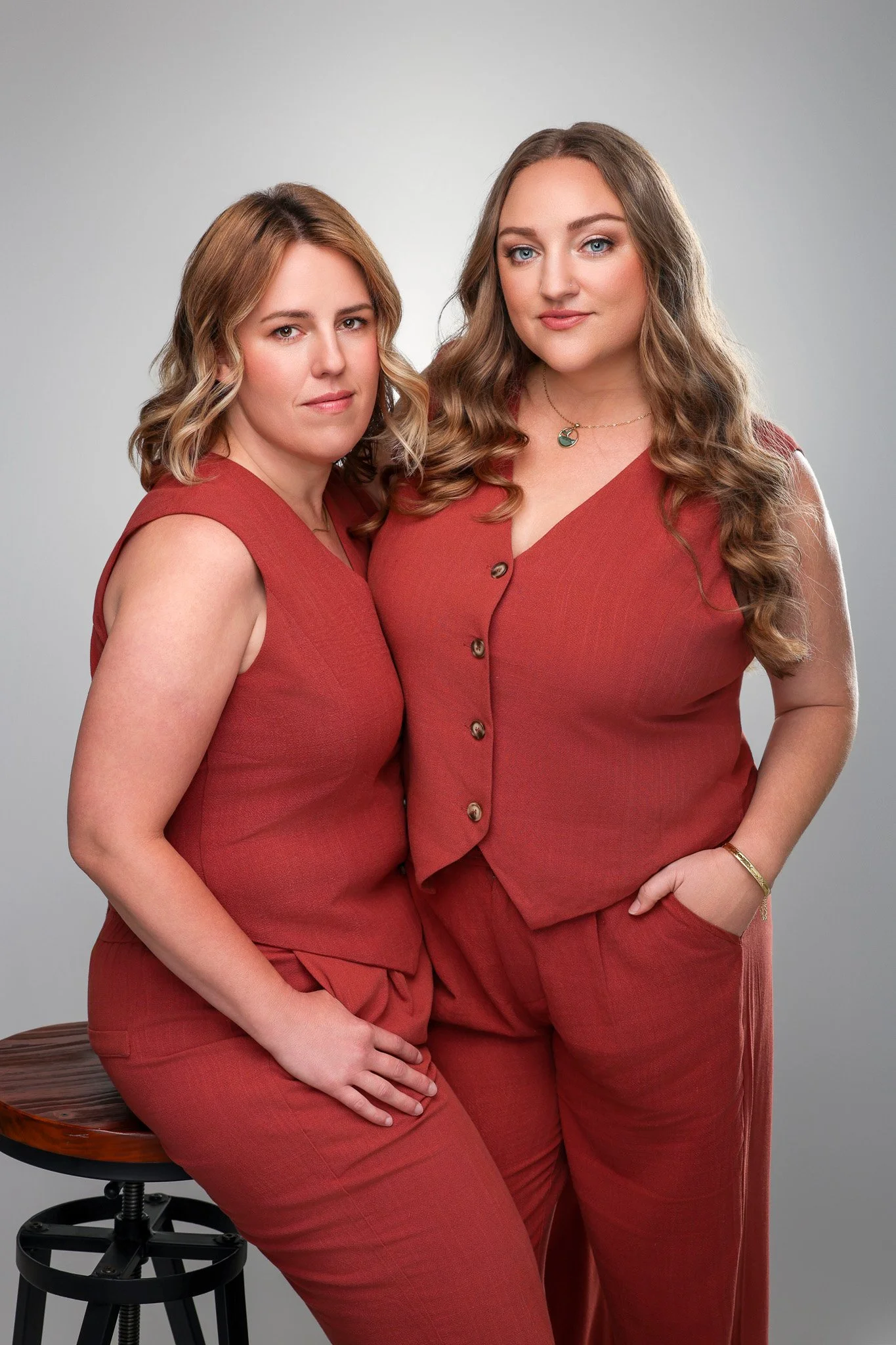 Two women with light skin and long, wavy hair in matching rust-colored outfits posing together in a studio. One seated on a stool and the other standing beside her.