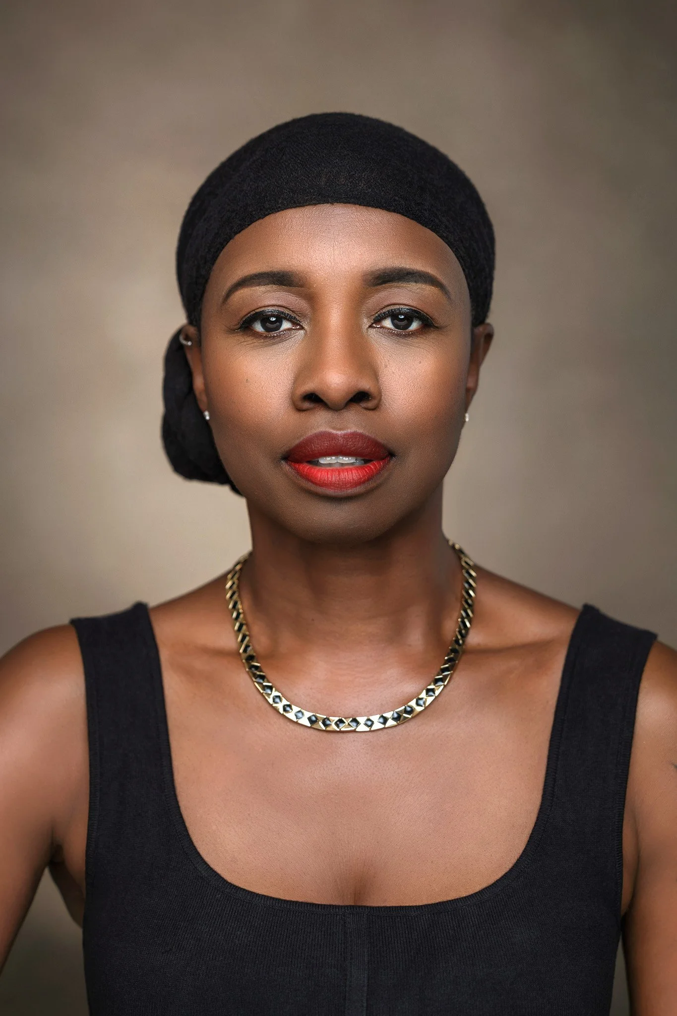 A woman wearing a black head wrap, black sleeveless top, and gold necklace with a neutral background.