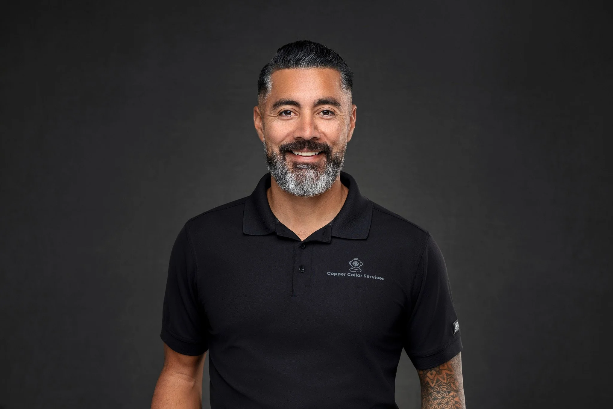 A smiling man with gray hair and a beard wearing a black polo shirt with a logo and text, standing against a dark background.