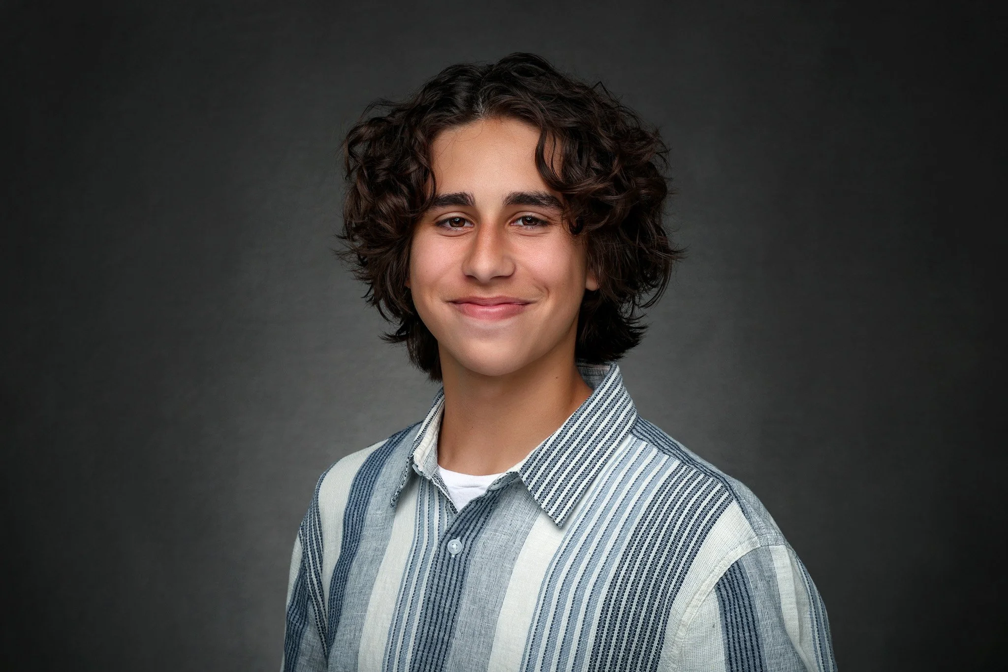 Portrait of a young man with curly brown hair, wearing a striped button-up shirt, smiling slightly against a dark background.