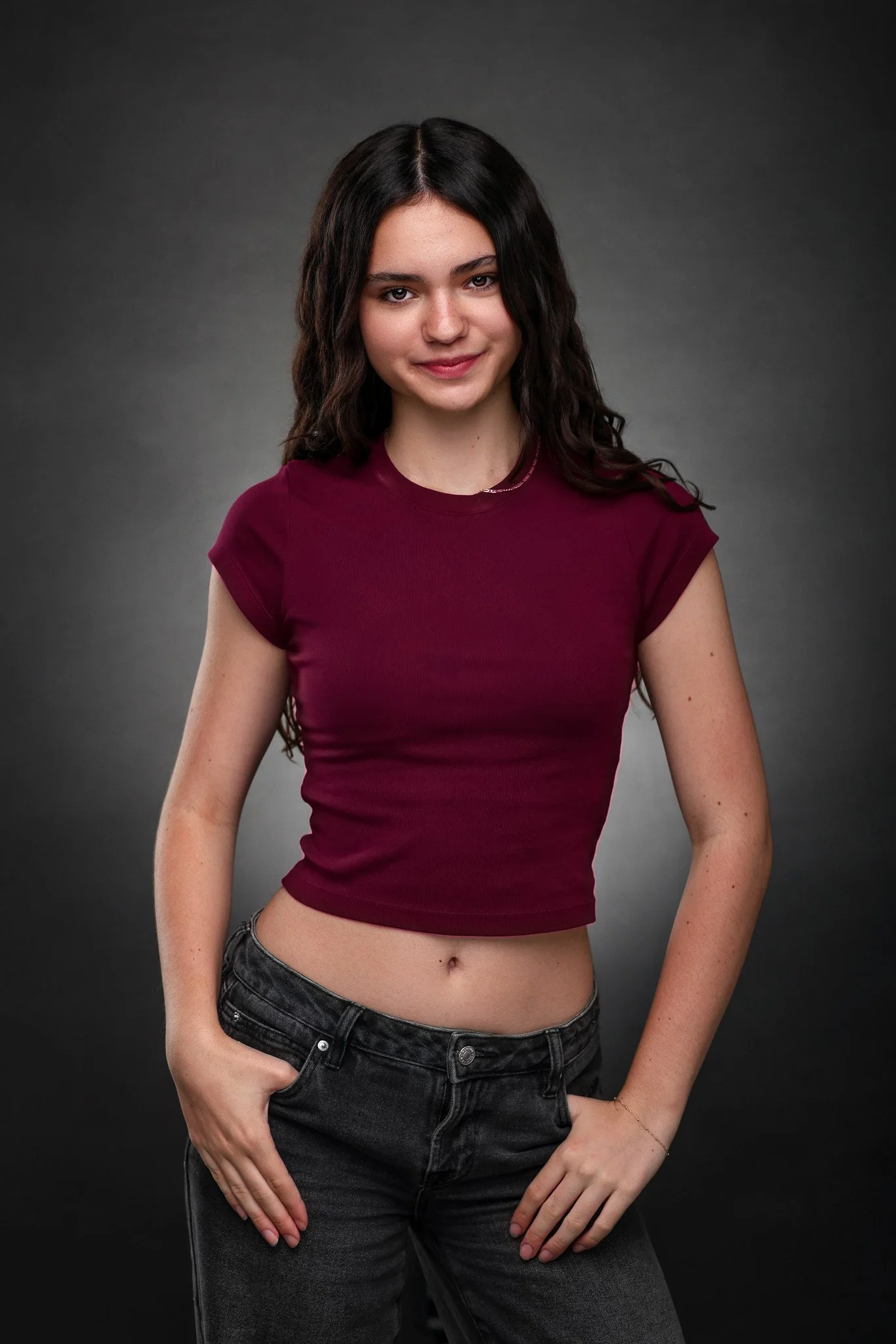 Young woman with dark brown hair in a wavy hairstyle, wearing a maroon cropped t-shirt and high-waisted black jeans, standing against a dark gray background.