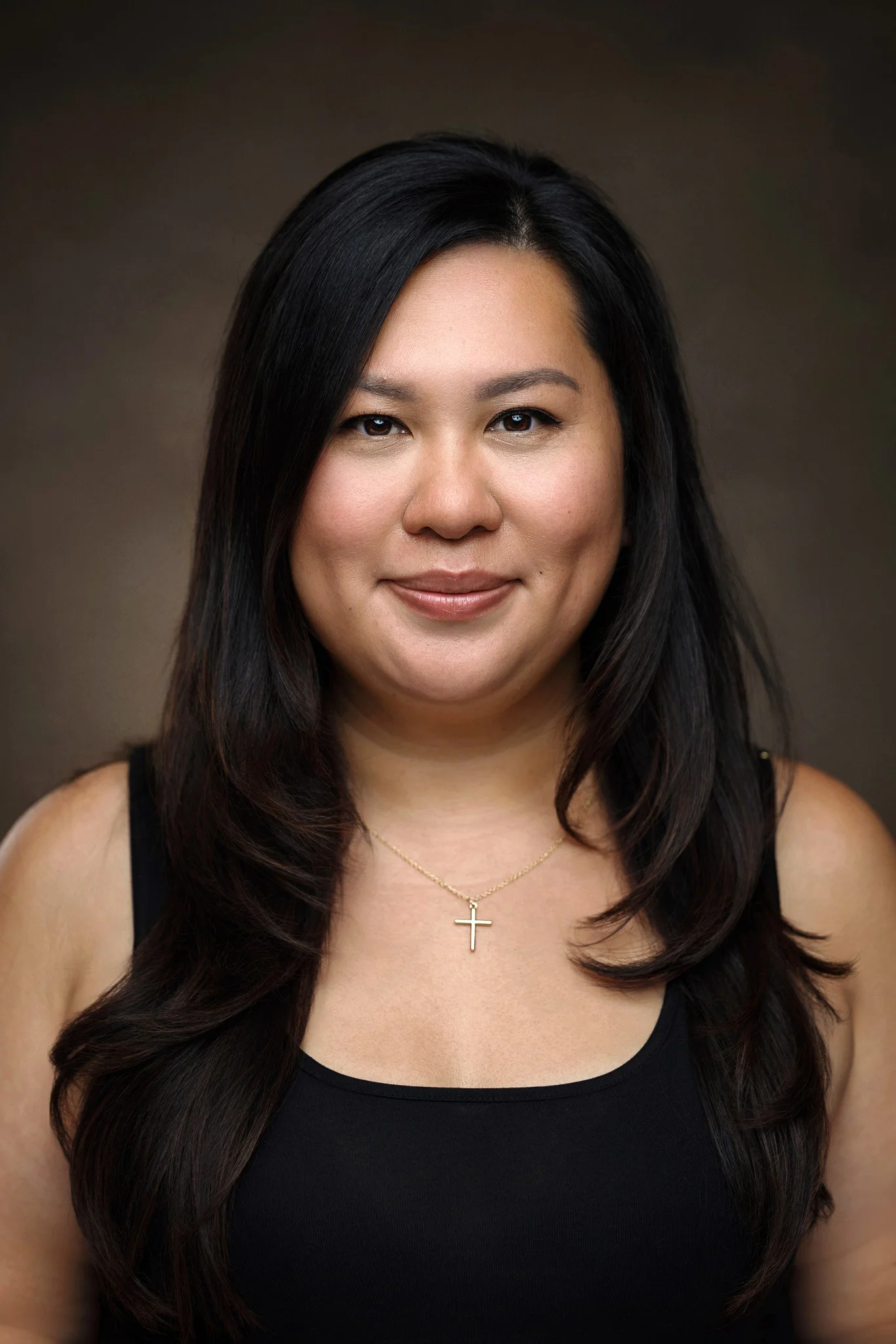 A woman with long black hair, wearing a black sleeveless top and a gold cross necklace, smiling at the camera against a brown background.