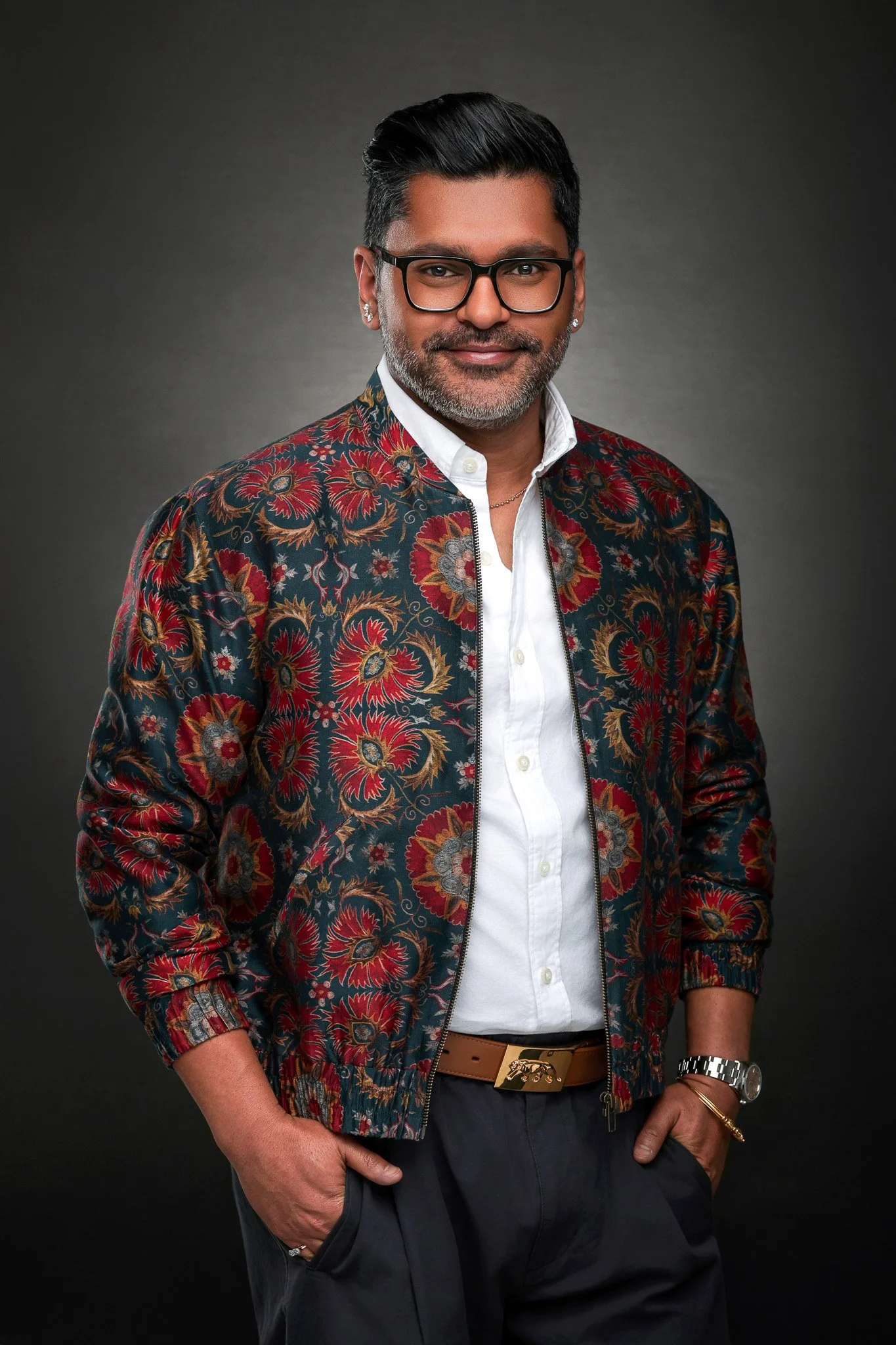 A man with glasses and a beard wearing a patterned jacket over a white shirt, with hands in pockets, standing against a dark gray background.