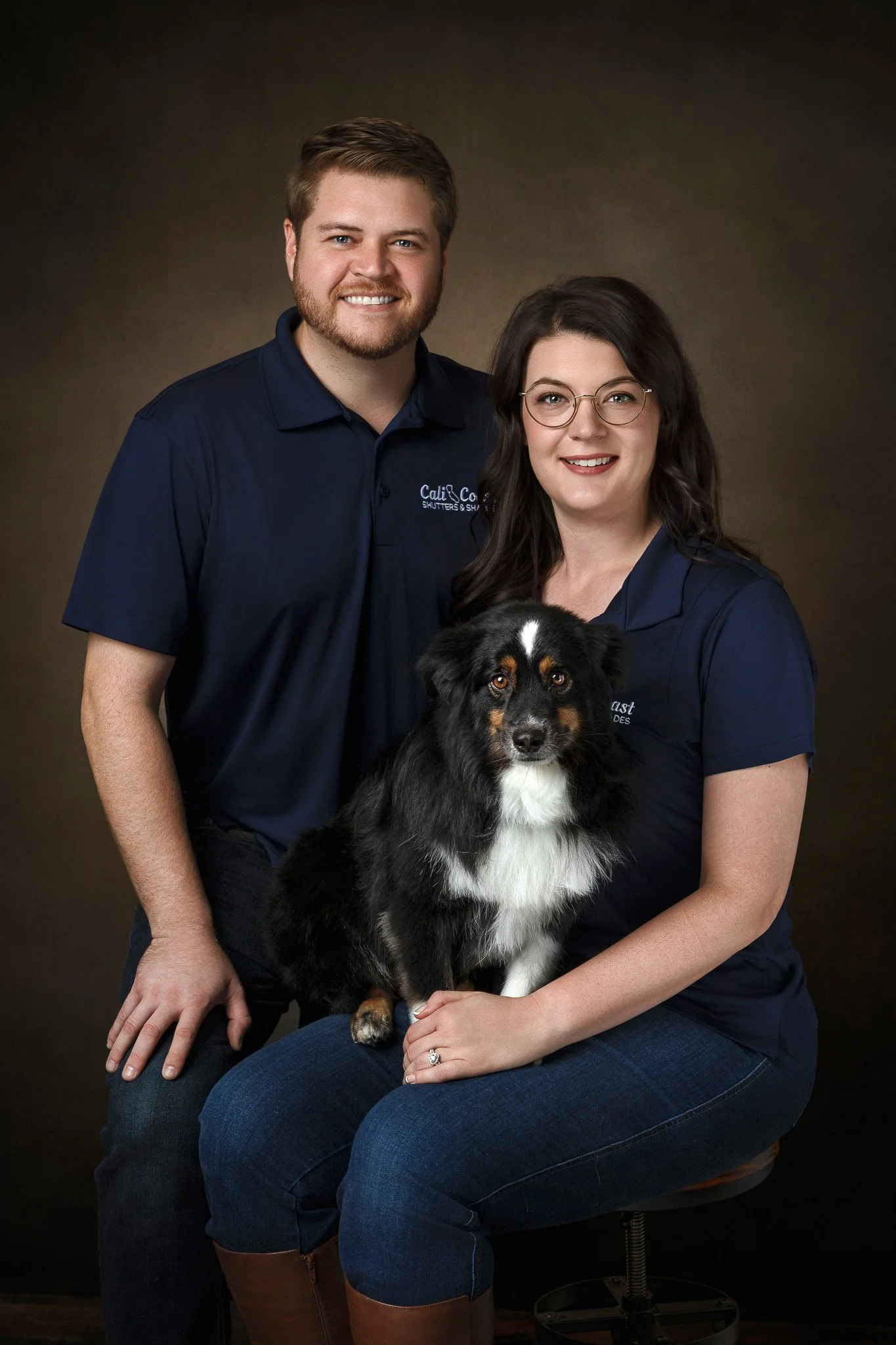 A woman with glasses sitting on a stool, holding a black, white, and brown Australian Shepherd dog on her lap. A man standing behind her, with his hand on her shoulder. They are all dressed in navy blue shirts, in a studio setting with a dark brown b