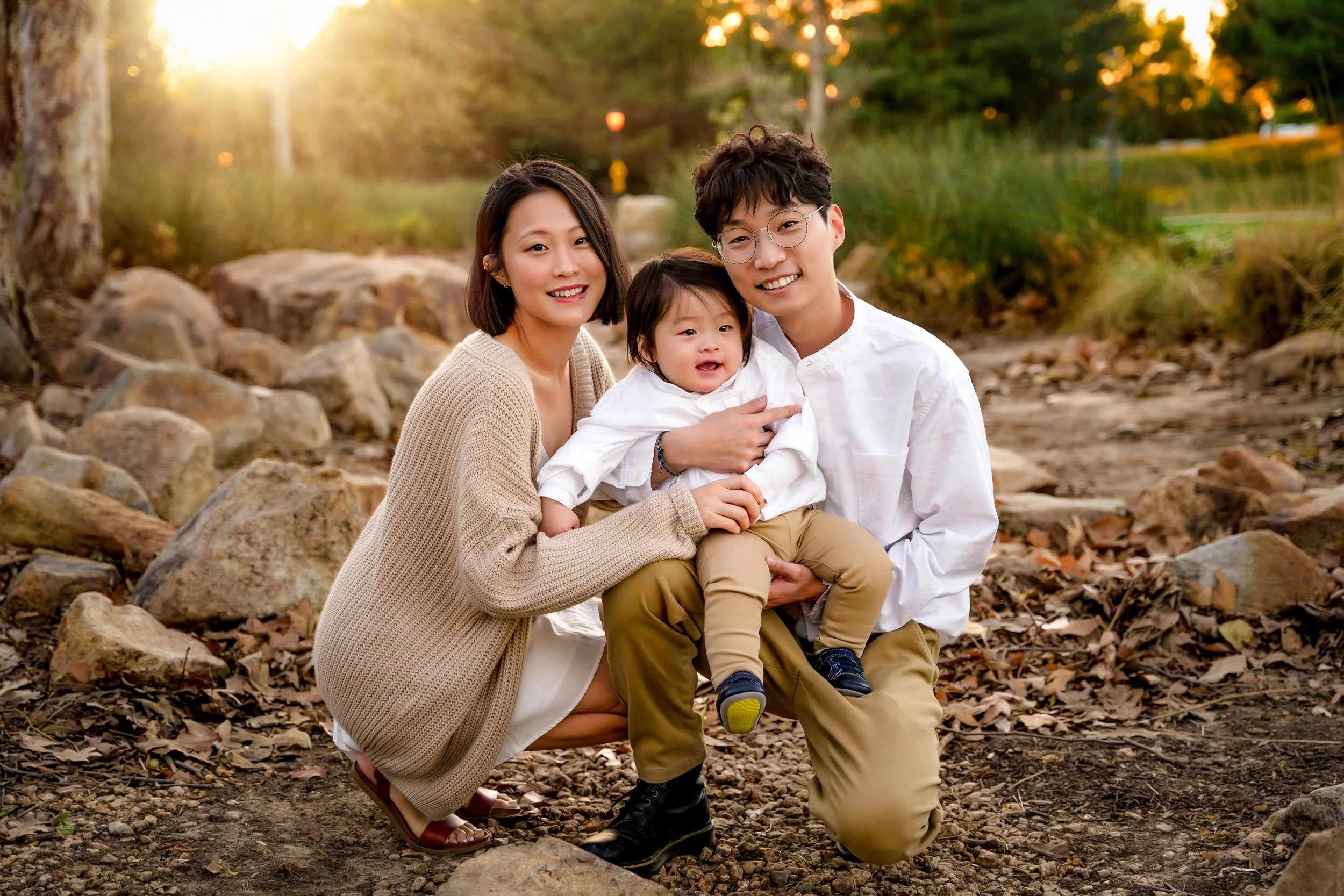 A family with a woman, a man, and a young child poses outdoors among rocks and trees at sunset, smiling at the camera.
