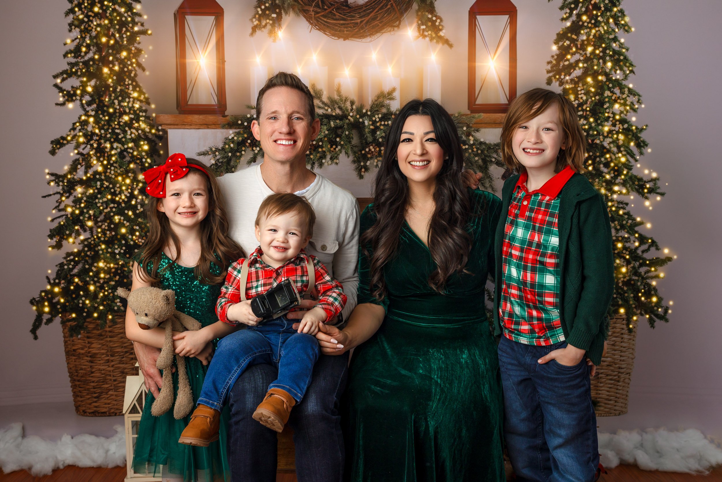 A family of six poses in front of decorated Christmas trees with lights, candles, and lanterns. The family includes two adults and four children, all dressed in festive holiday clothing, smiling for the photo.