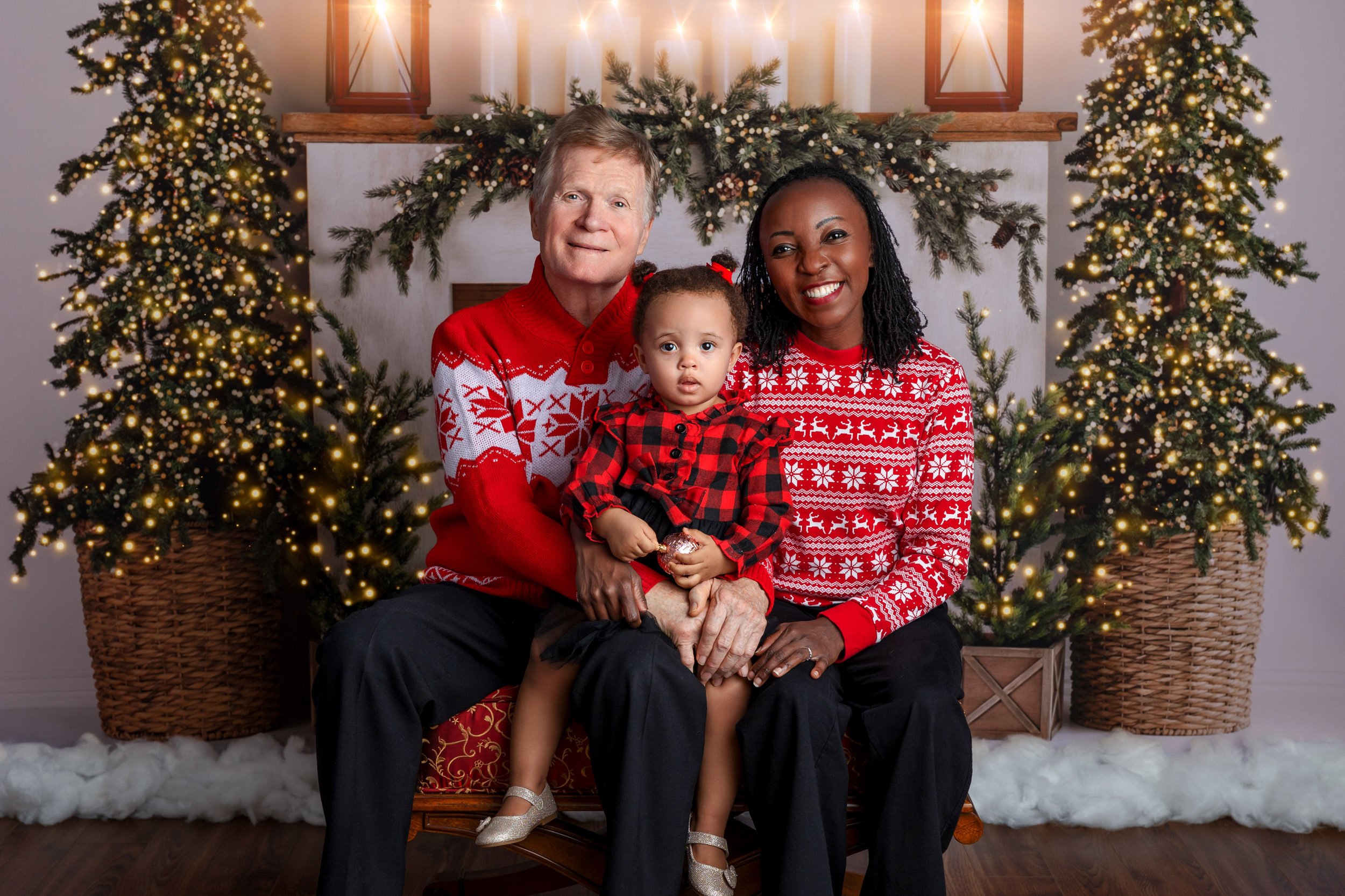 Three family members, a man, woman, and young girl, sitting in front of Christmas decorated fireplace with lit Christmas trees, wearing matching red and white holiday sweaters.