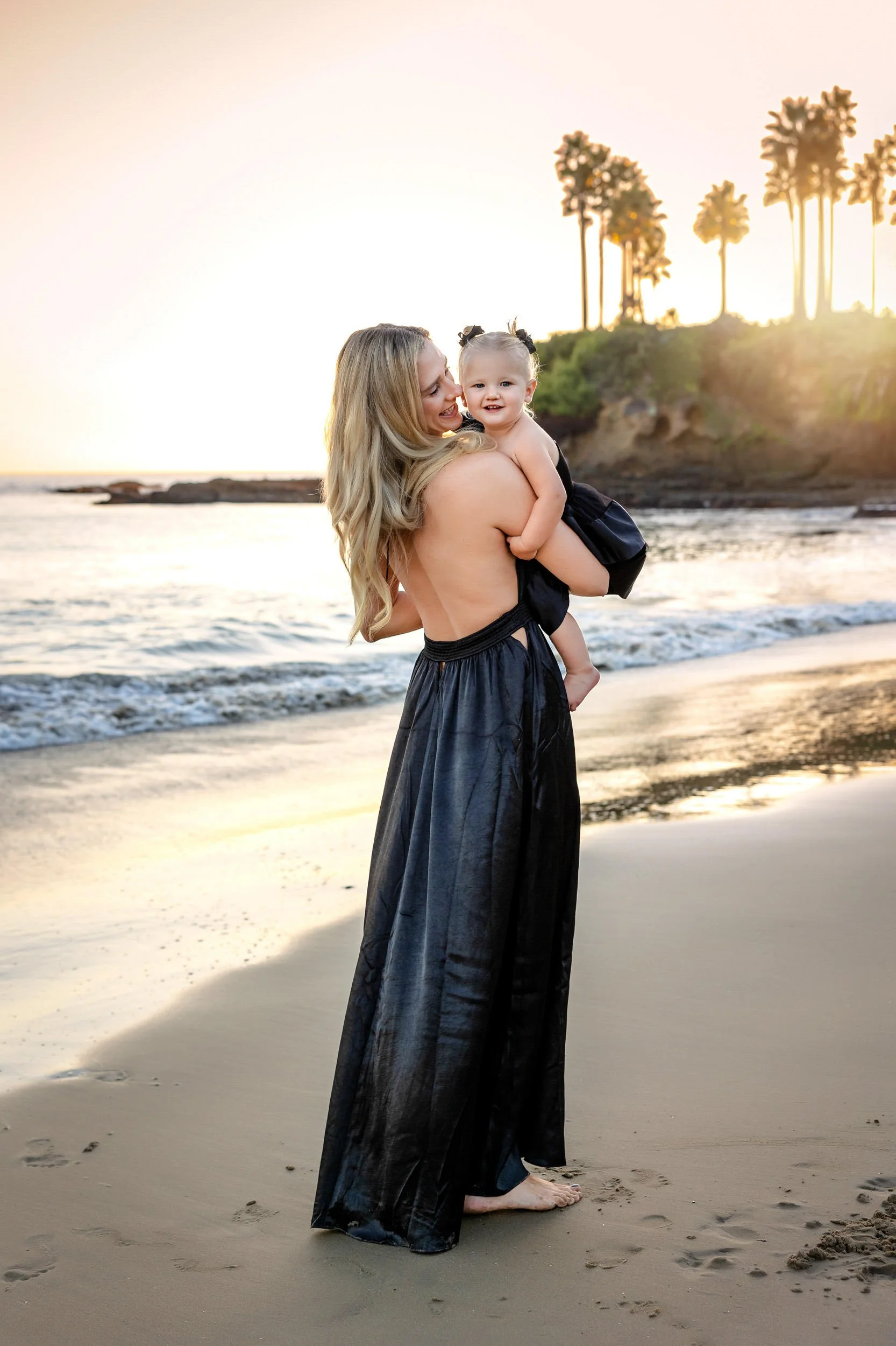 A woman in a long black dress holding a child on a beach at sunset, with palm trees in the background.