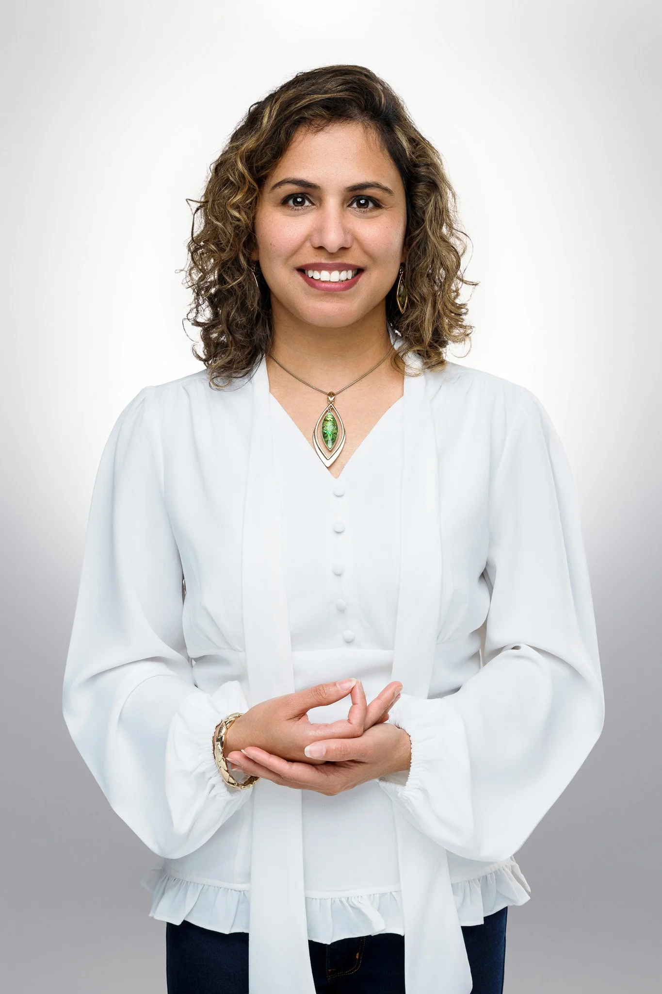A woman with curly brown hair, wearing a white blouse, smiling, standing against a neutral background, with jewelry including a green pendant necklace and earrings.