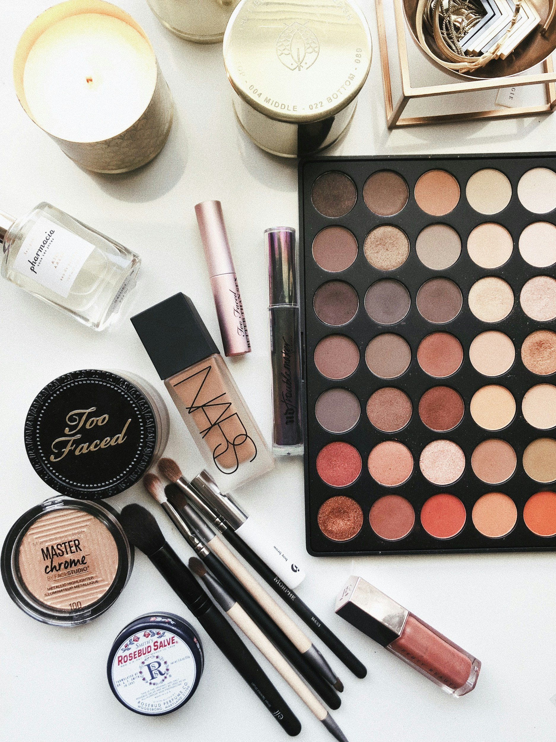 Makeup tools and products on a white surface, including an eyeshadow palette, brushes, foundation, lipstick, blush, and candles.