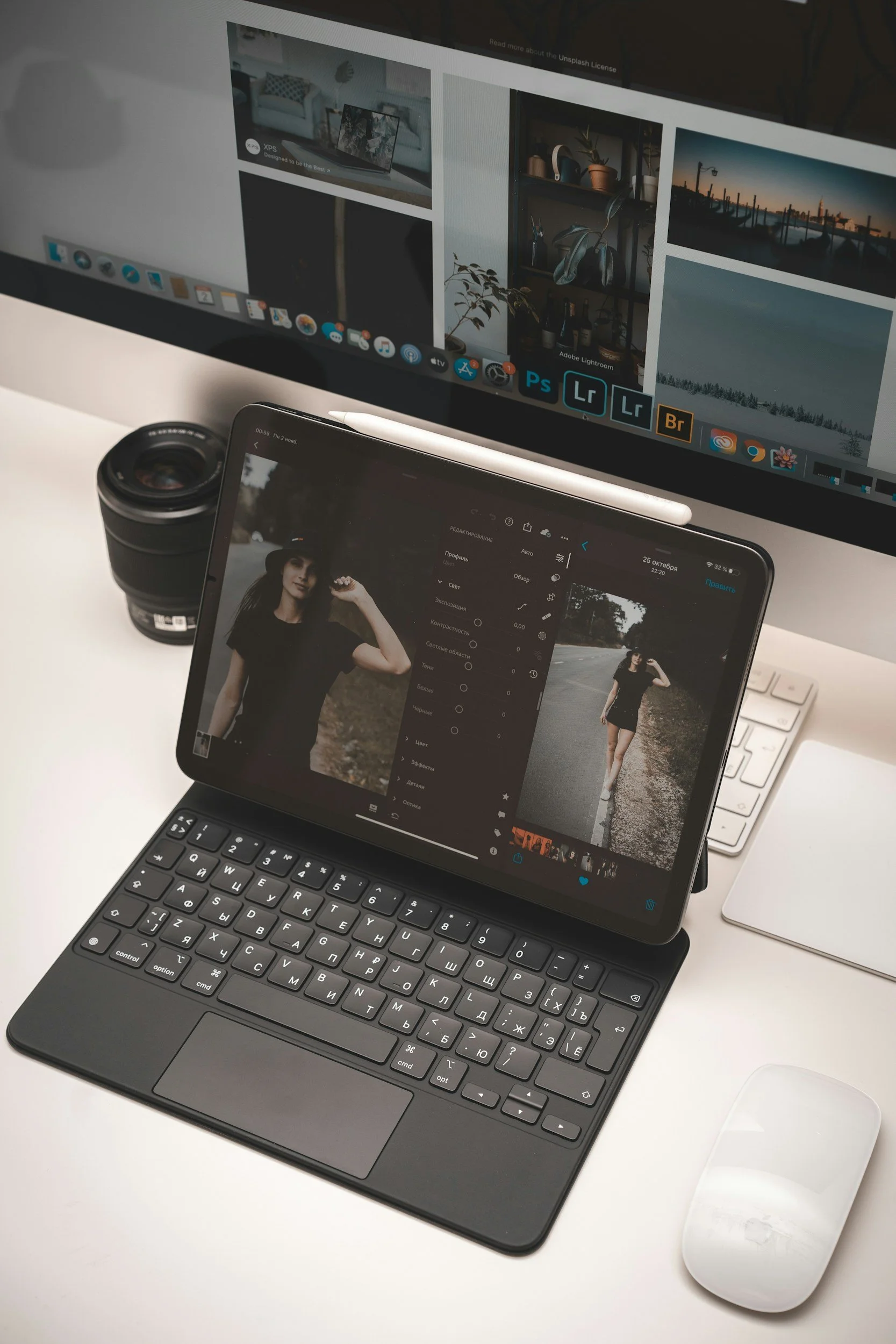 A workspace with a computer monitor displaying photo editing software, a tablet with a photo of a woman in black, a camera lens, a wireless keyboard, and a mouse on a white desk.