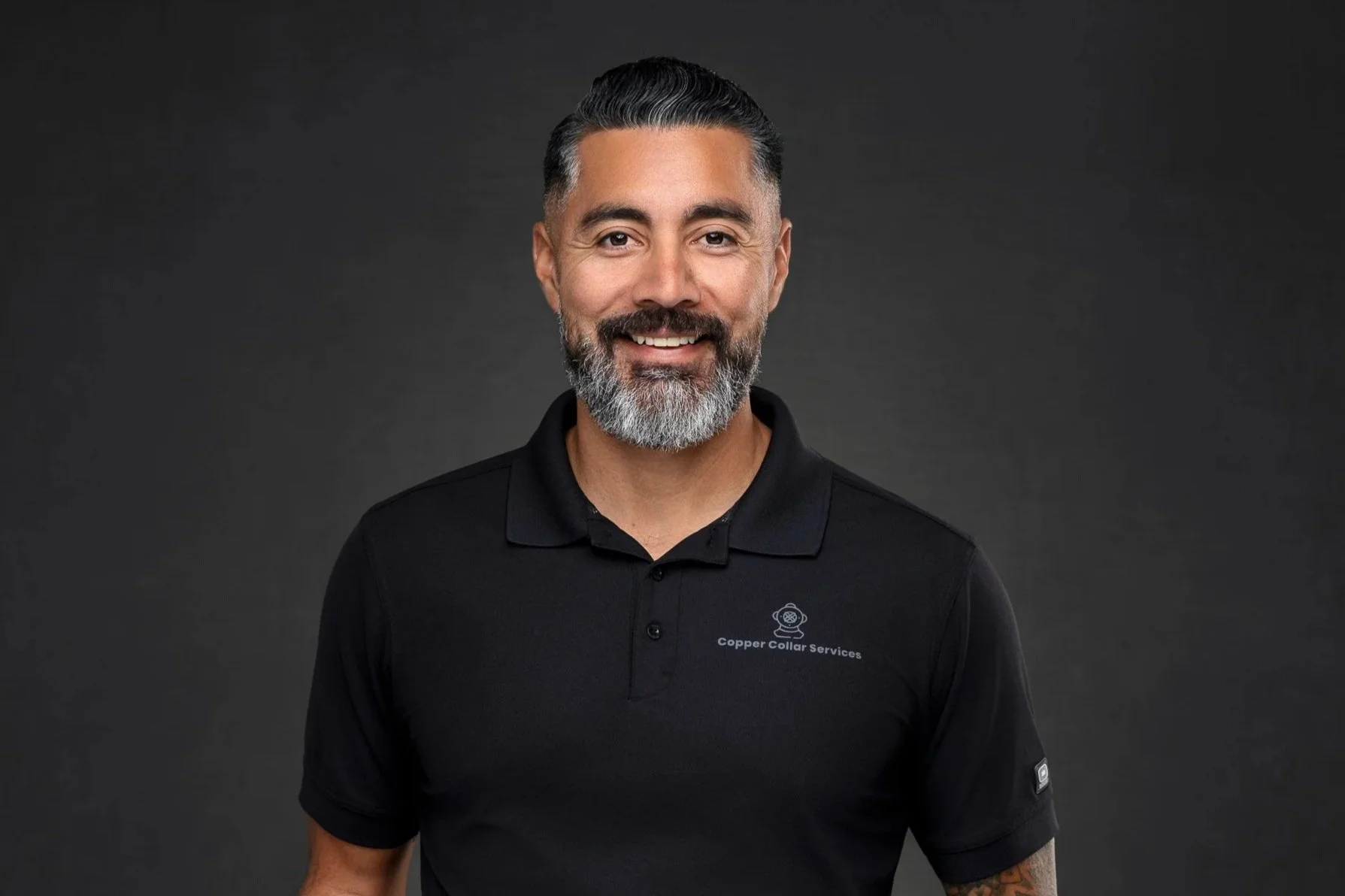 Smiling man with gray beard and dark hair, wearing a black polo shirt with a logo and text that says 'Copper Collar Services', standing against a dark gray background.