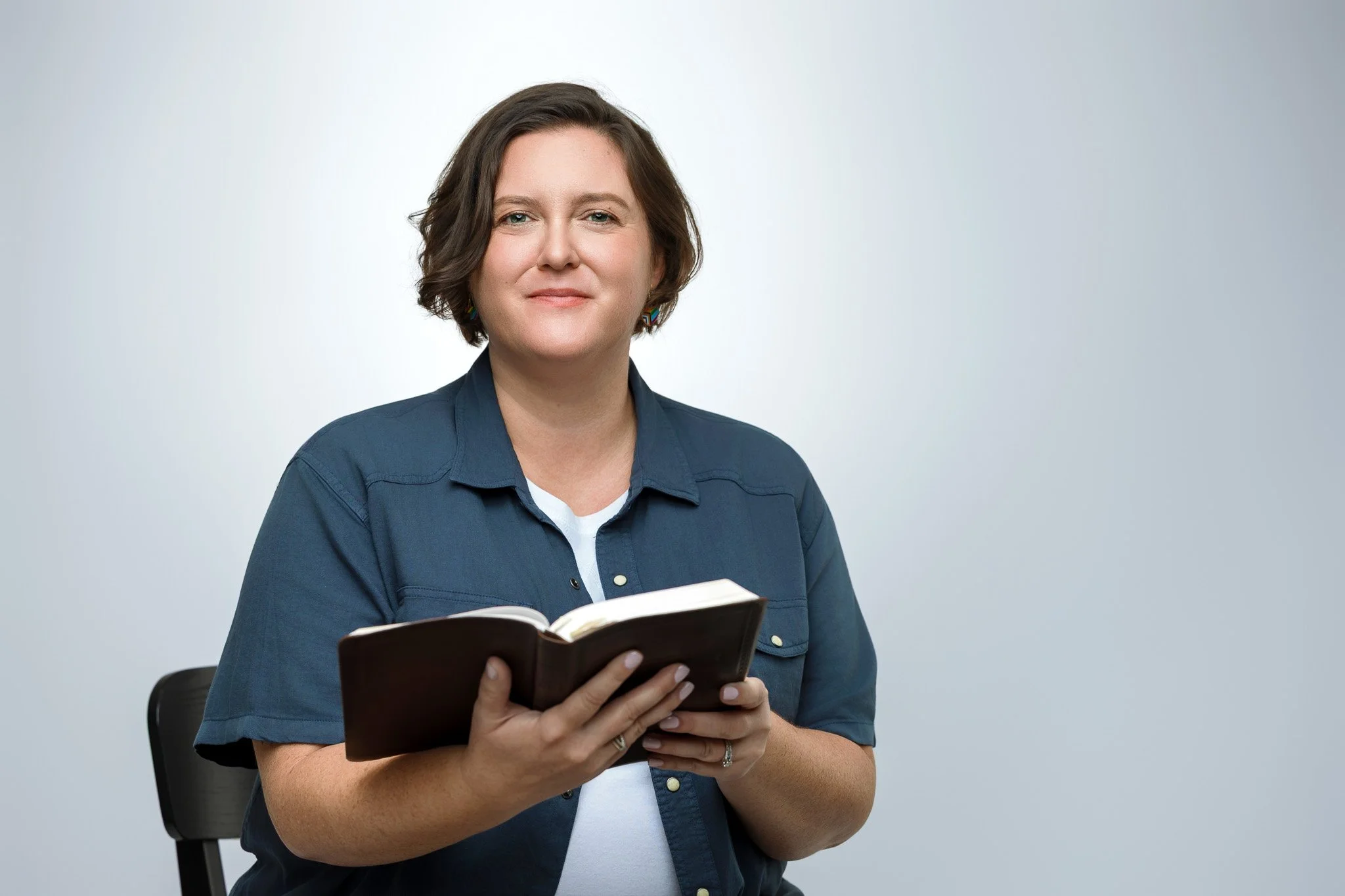 A woman with short brown hair smiling while holding an open book, sitting on a black chair against a plain light gray background.