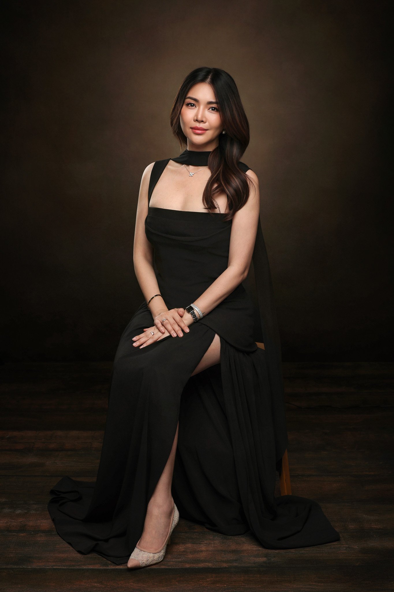 A woman with long dark hair sitting in a black dress with a high slit, showing a beige high heel. She is seated on a wooden floor against a dark brown background.