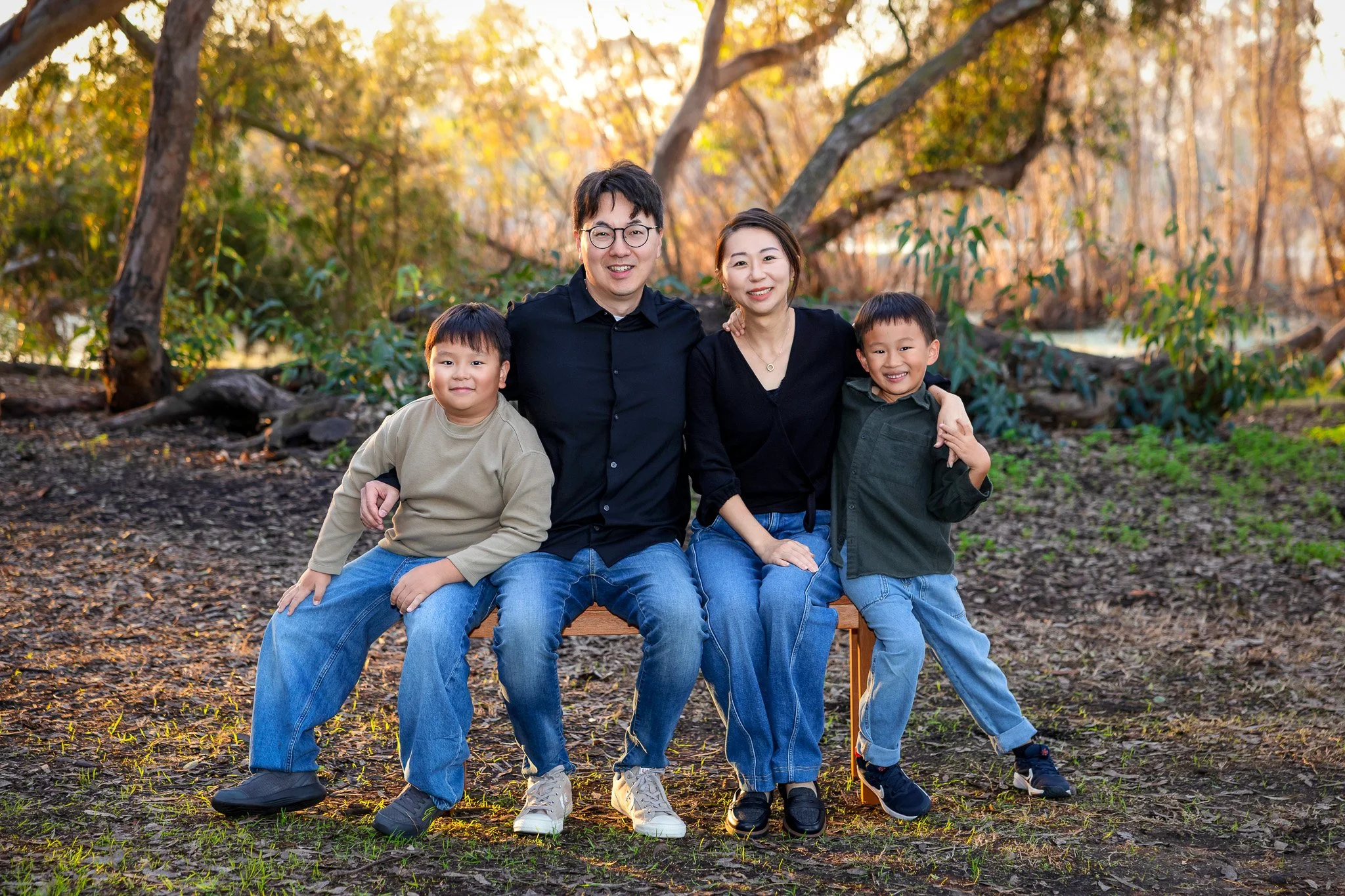 A happy family of four sitting on a bench outdoors in a park with autumn trees in the background, smiling at the camera.