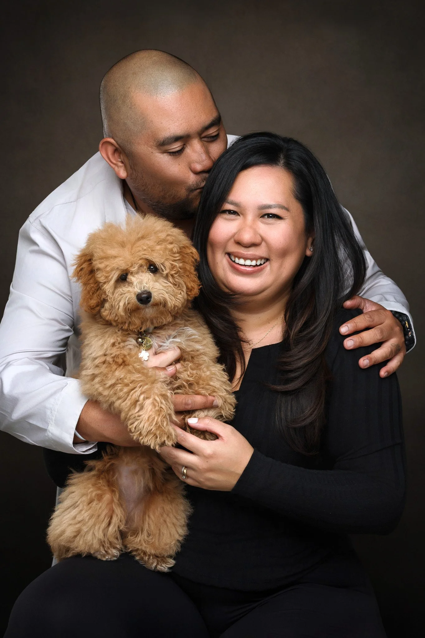 A man kissing a woman on the forehead while holding a small, fluffy, tan poodle. The woman is smiling and sitting down, with the dog in her lap against a dark background.