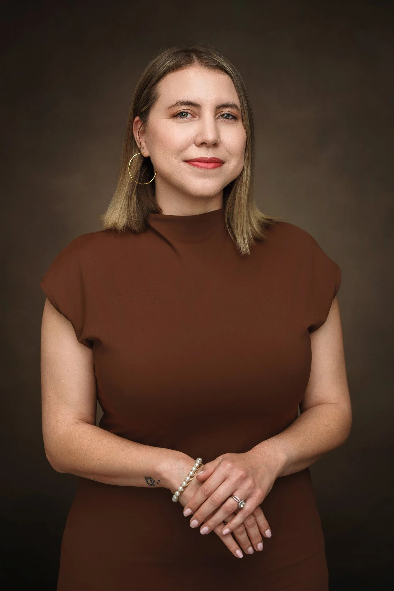 Front-facing portrait of a woman with shoulder-length light brown hair, blue eyes, and light skin, wearing a brown top, gold hoop earrings, a pearl bracelet, and a ring, standing against a dark brown background.