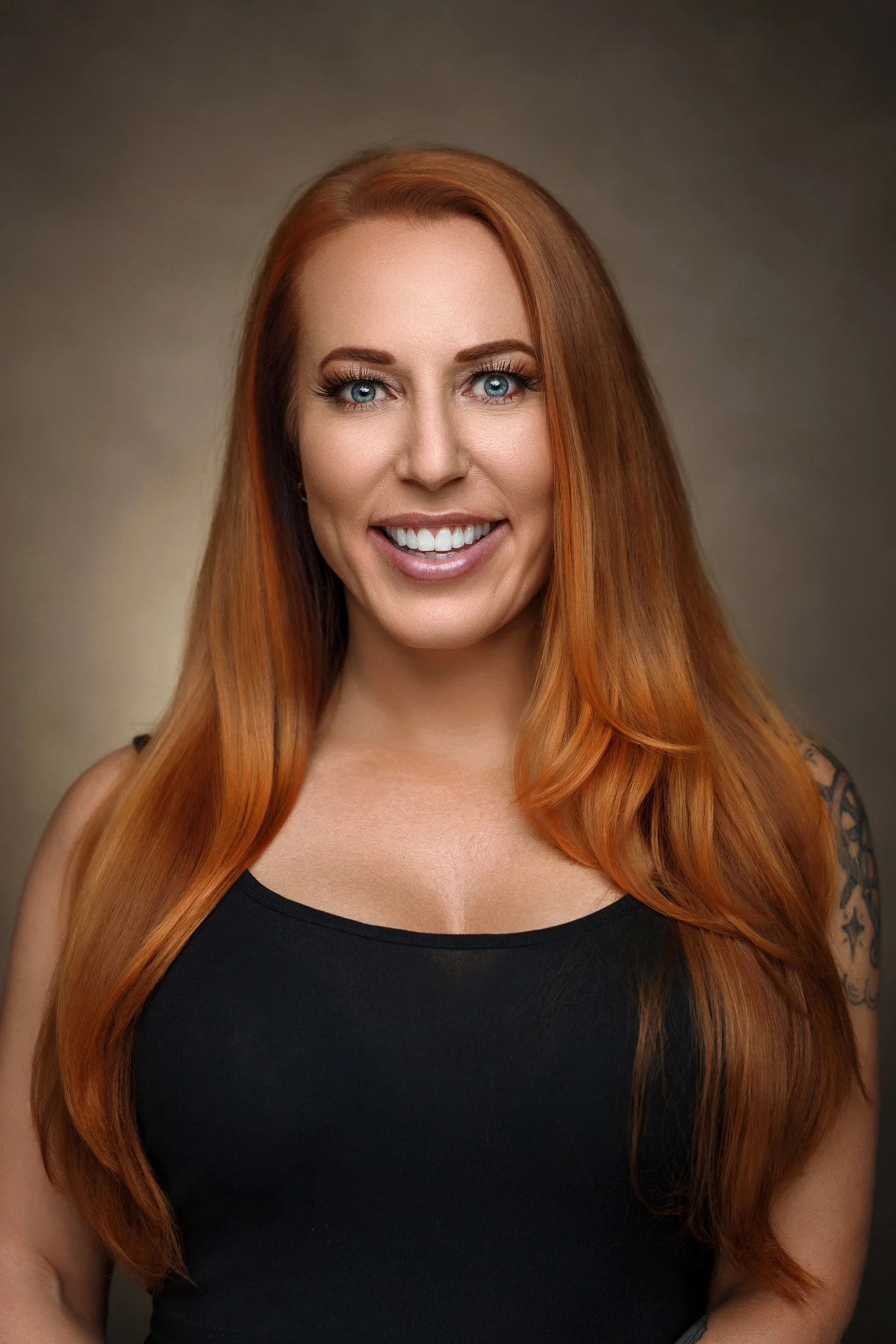 A woman with long red hair and blue eyes smiling, wearing a black sleeveless top, with tattoos on her arm, against a neutral background.