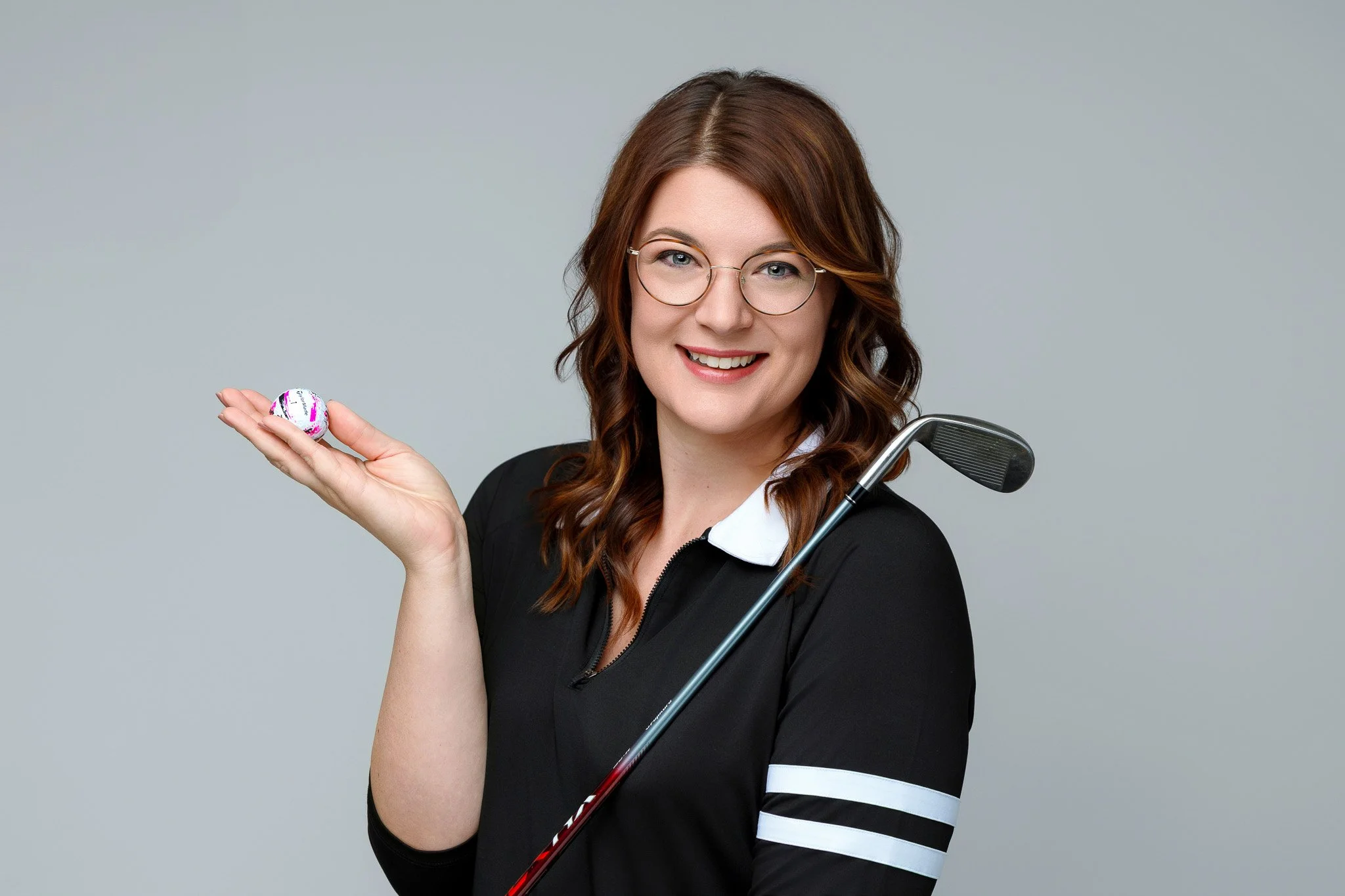 A young woman with glasses and long, wavy brown hair smiling, holding a golf ball in her left hand, and gripping a golf club with her right arm, wearing a black jacket with white stripes on the sleeves, against a plain gray background.