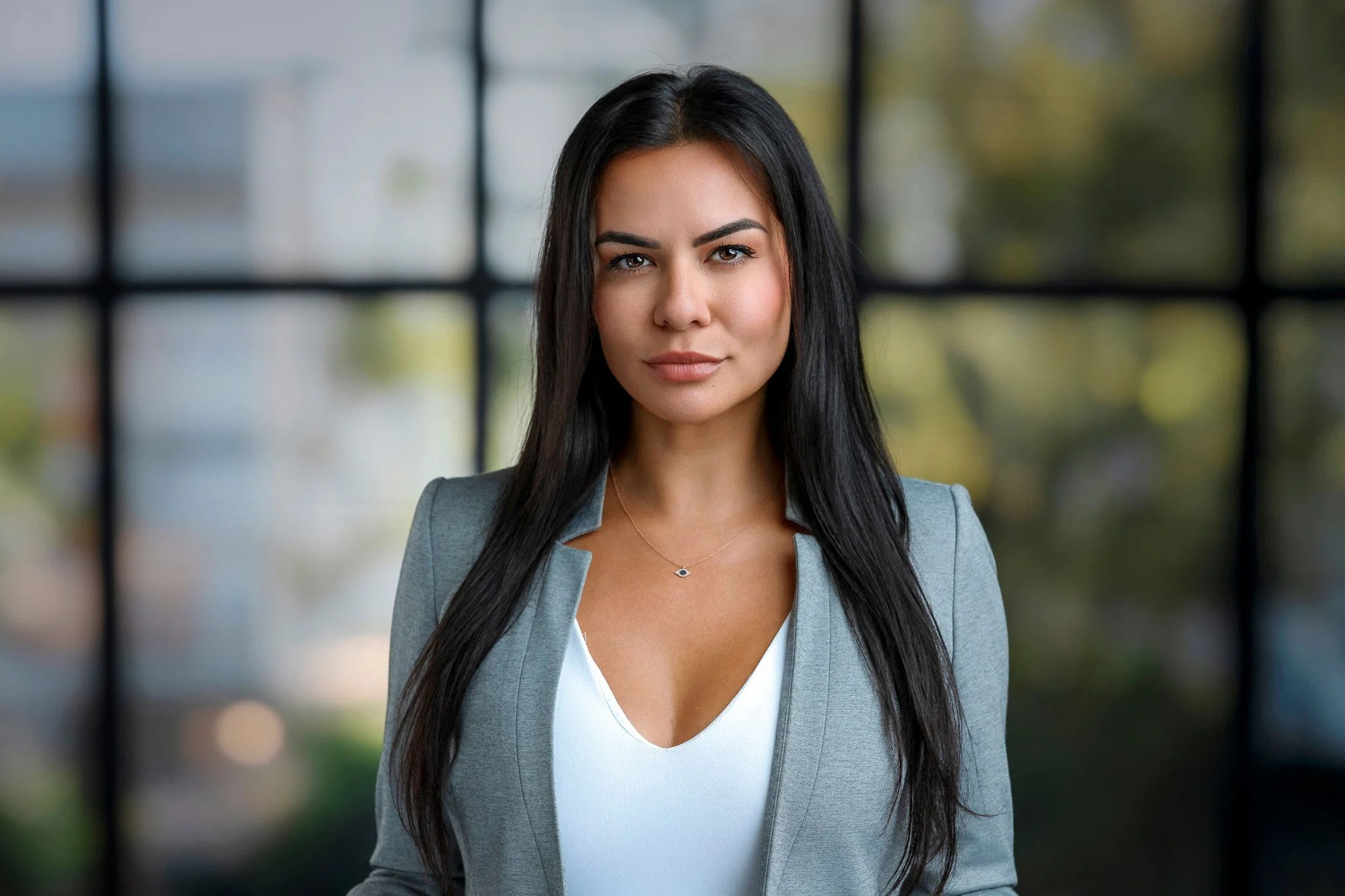 A woman with long dark hair, wearing a gray blazer and white top, standing in front of a large window with blurred outdoor scenery.