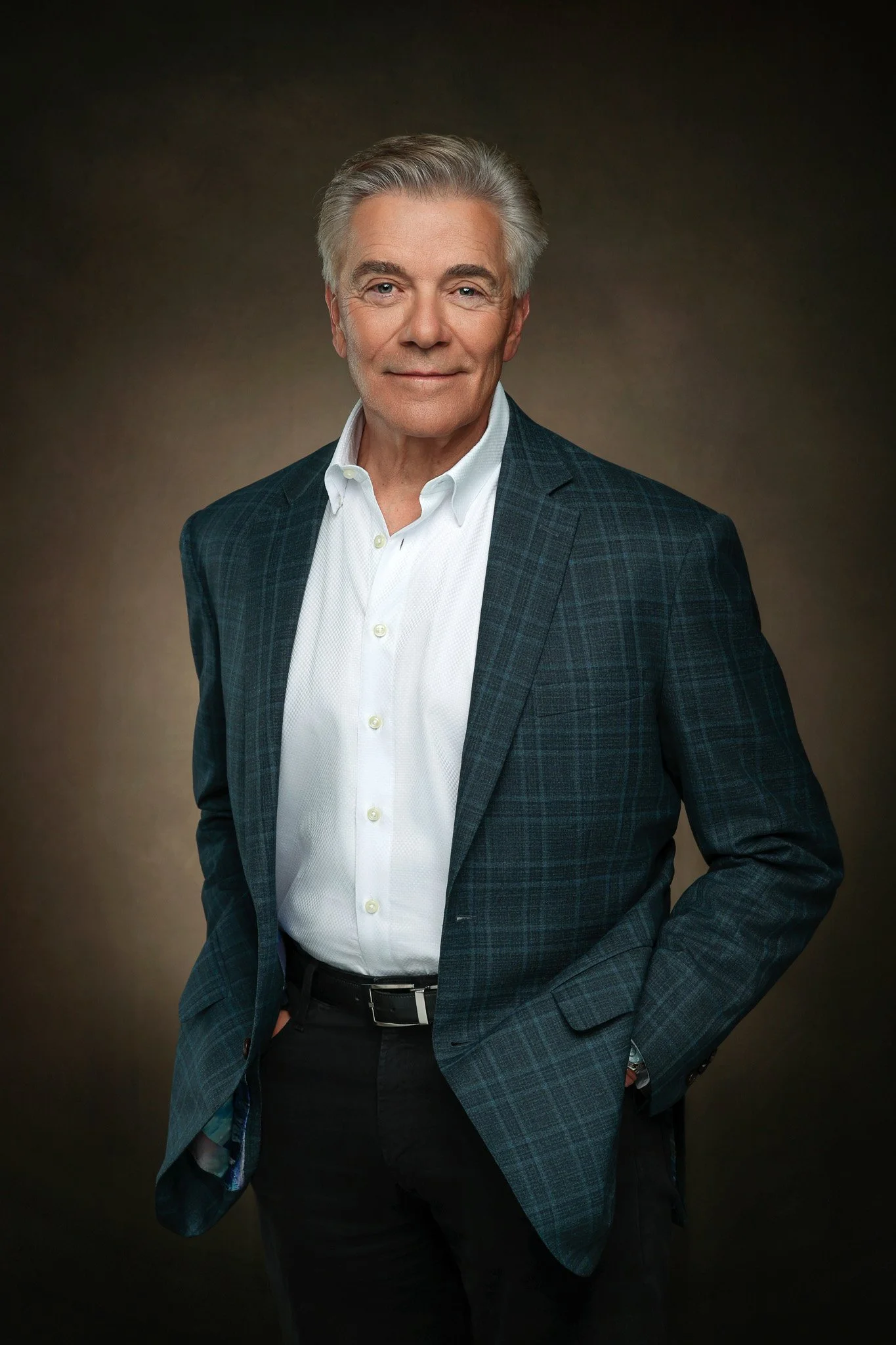 Portrait of an older man with gray hair wearing a white shirt and a dark checkered blazer, standing with one hand in his pocket against a gradient brown background.