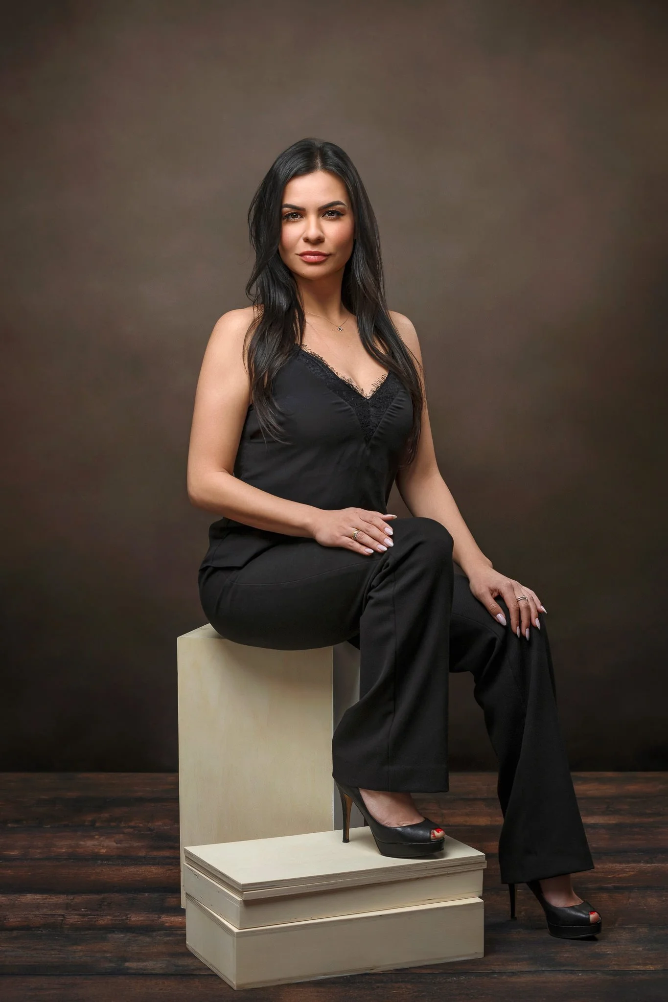 A woman with long dark hair in a black sleeveless top and black pants sitting on a white block against a brown gradient background.