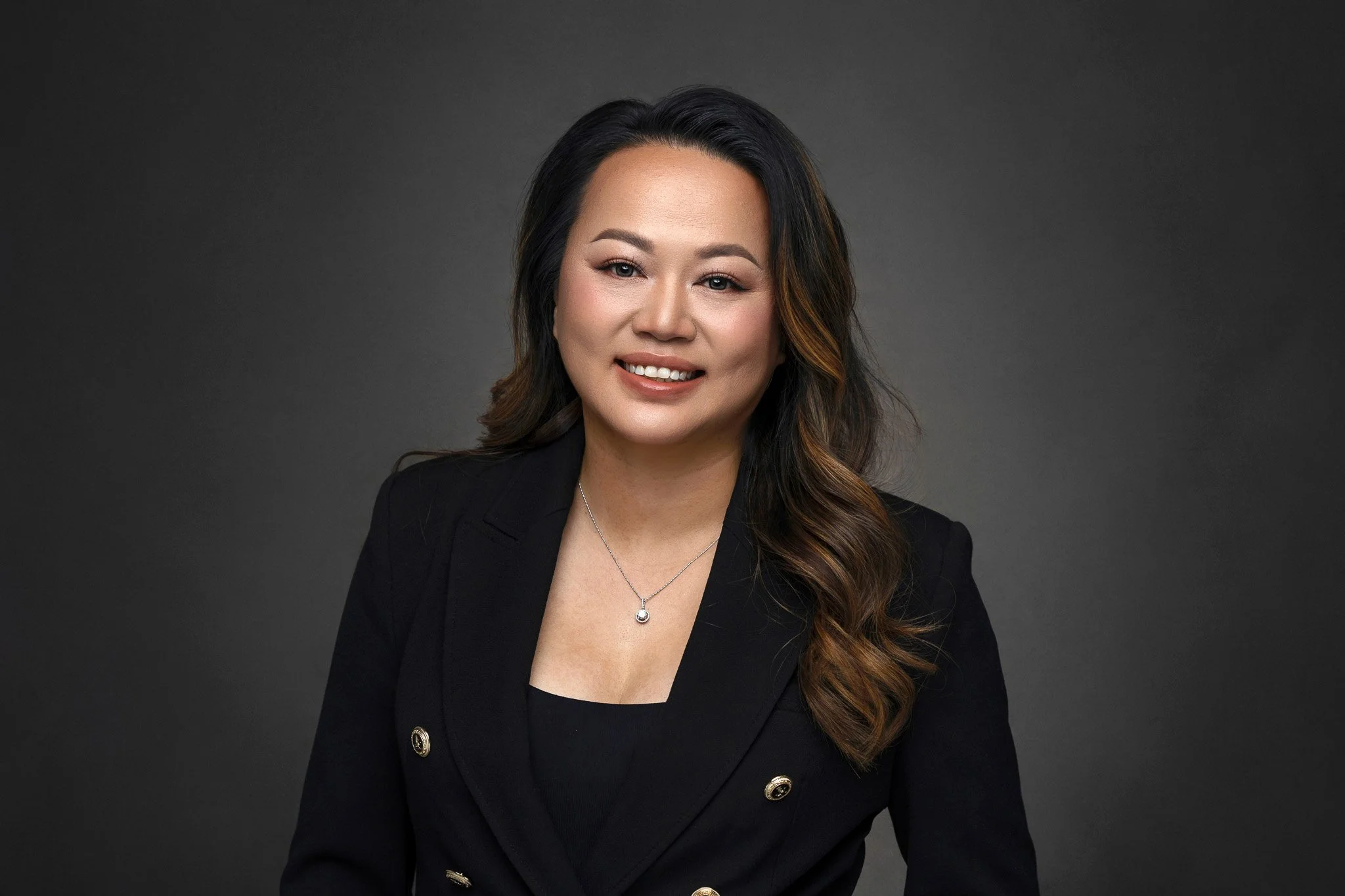 A woman with long dark hair with brown highlights, wearing a black blazer with gold buttons, a black top, and a silver necklace with a pendant, smiling against a dark gray background.