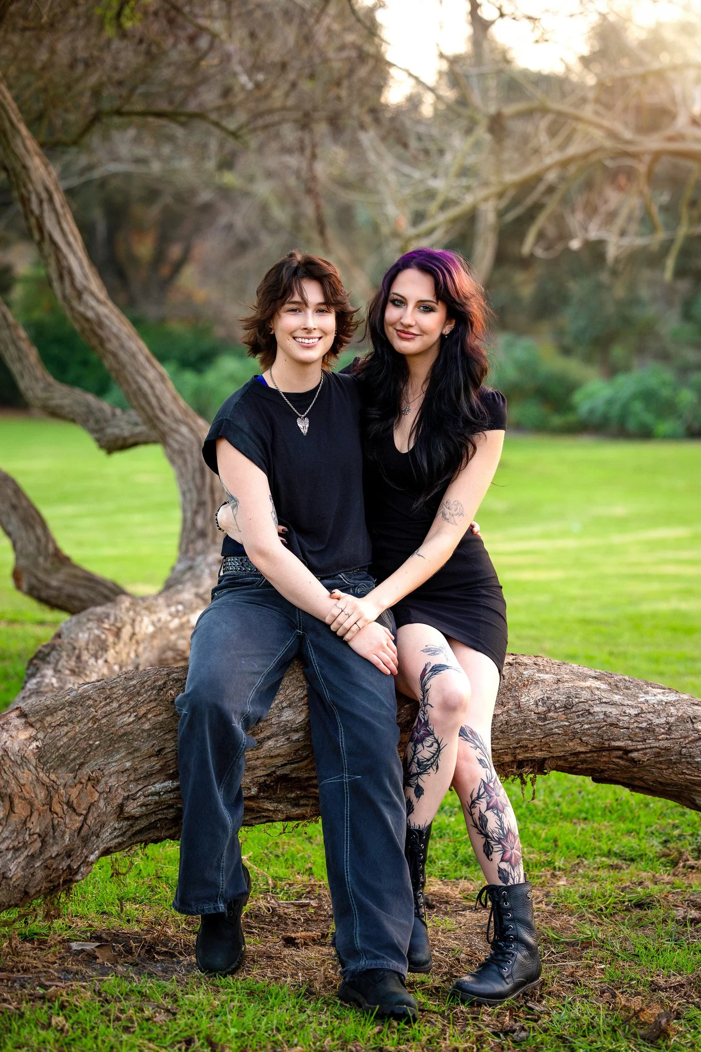 Two women sitting closely on a large tree branch in a park during sunset, smiling at the camera. One has short brown hair, wears a black t-shirt and jeans. The other has long black hair, wears a black dress, tattoos on her legs, and black boots.