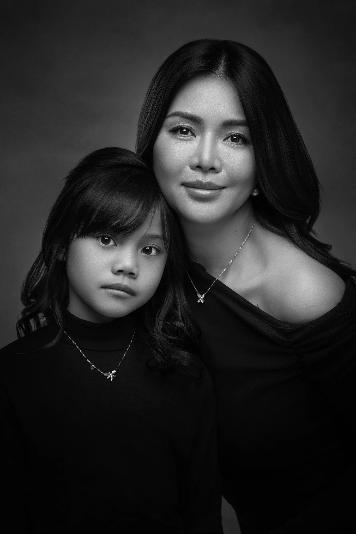 Black and white portrait of a woman and a girl, both wearing dark clothing and matching necklaces, with the woman positioned slightly behind and to the right of the girl, both looking directly at the camera.