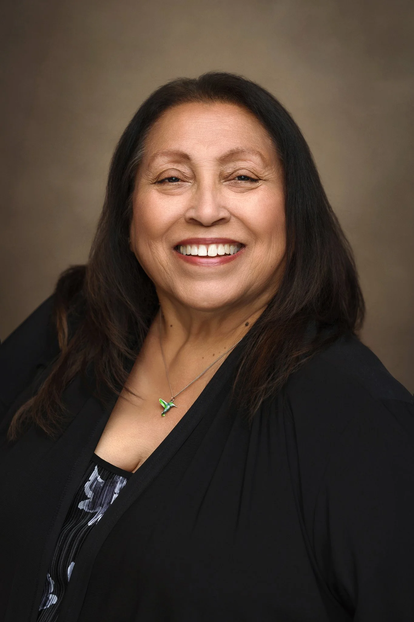 A smiling woman with long dark hair wearing a black blazer and a hummingbird necklace against a neutral background.