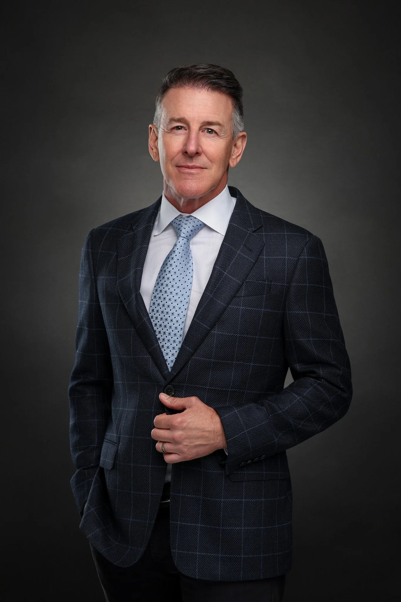 Portrait of a middle-aged man in a dark plaid suit, white shirt, and light blue tie, standing against a gray background.