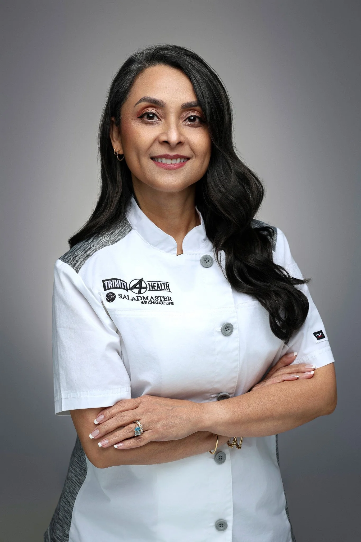 A woman in a white medical coat with Trinity Health logo, smiling with arms crossed, against a gray background.
