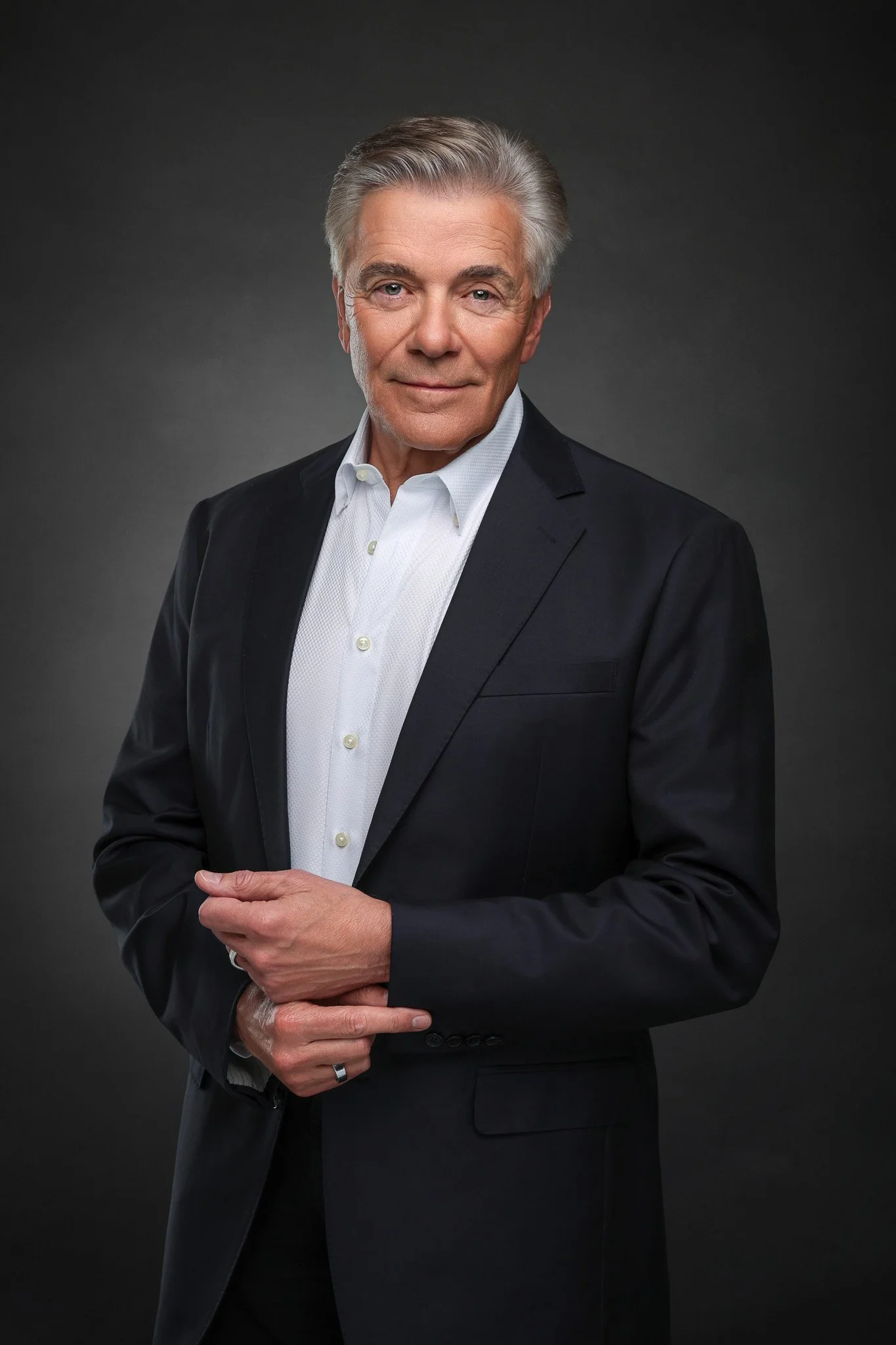An elderly man with gray hair wearing a black suit and white shirt, posed against a dark gray background.