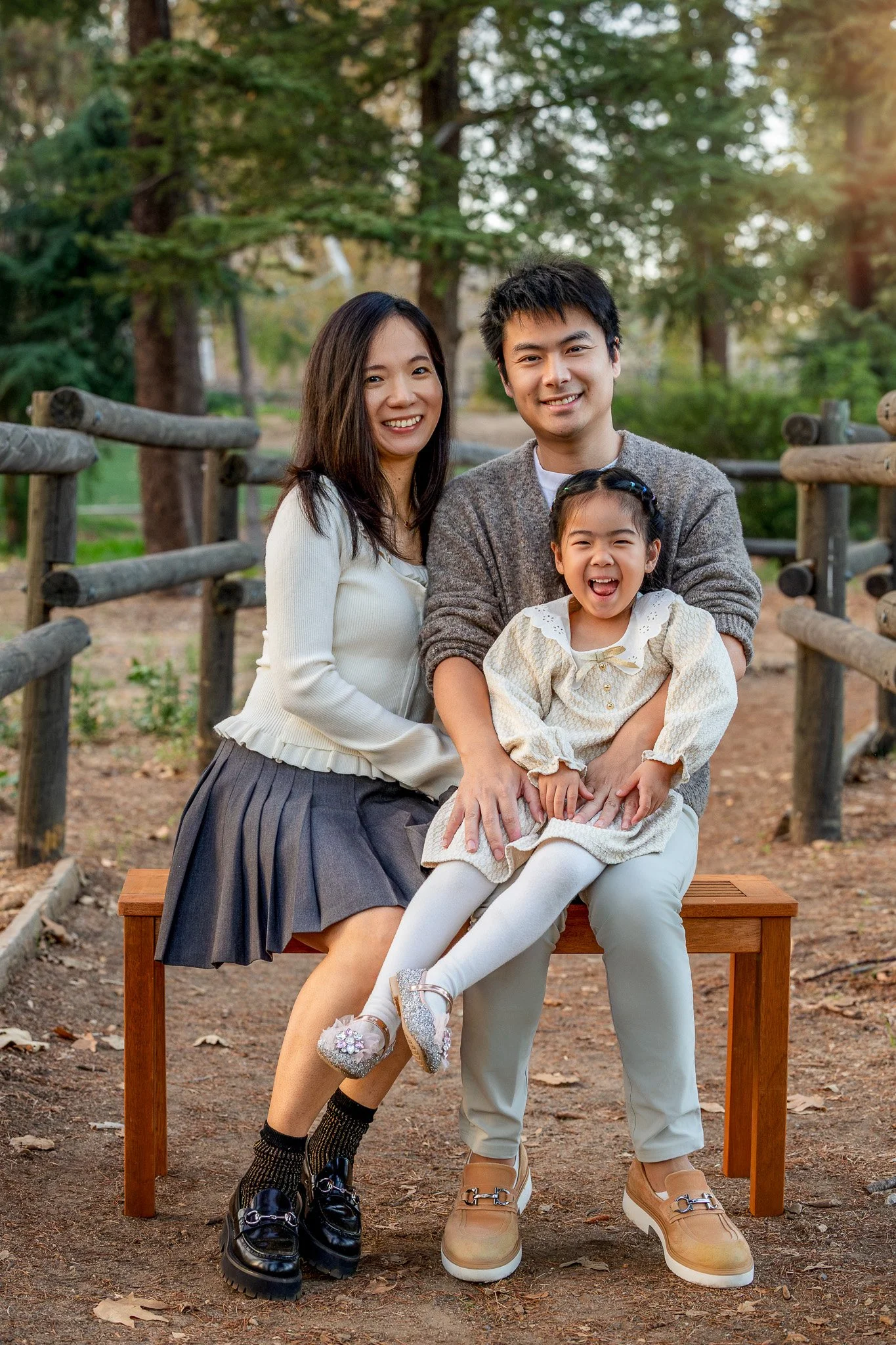 A family of three smiling and sitting on a wooden bench outdoors in a park with trees in the background.