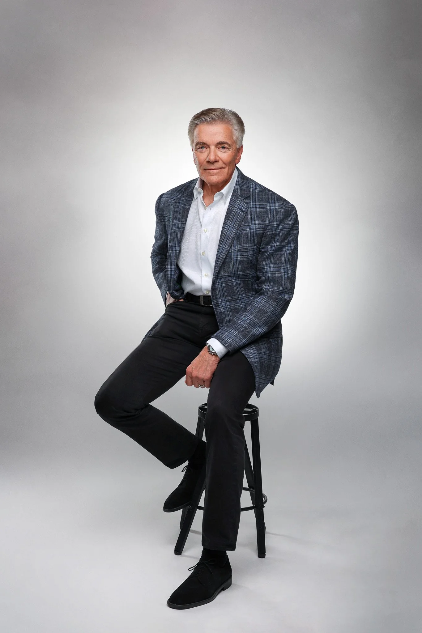 A mature man with gray hair and a confident expression, sitting on a black stool against a plain, light-colored background. He is dressed in a white shirt, a blue patterned blazer, and black pants.