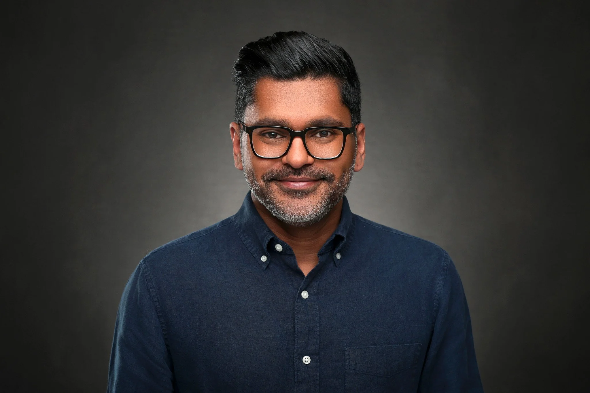 A middle-aged man with dark hair and a beard, wearing glasses and a navy blue button-up shirt, smiling at the camera against a dark background.
