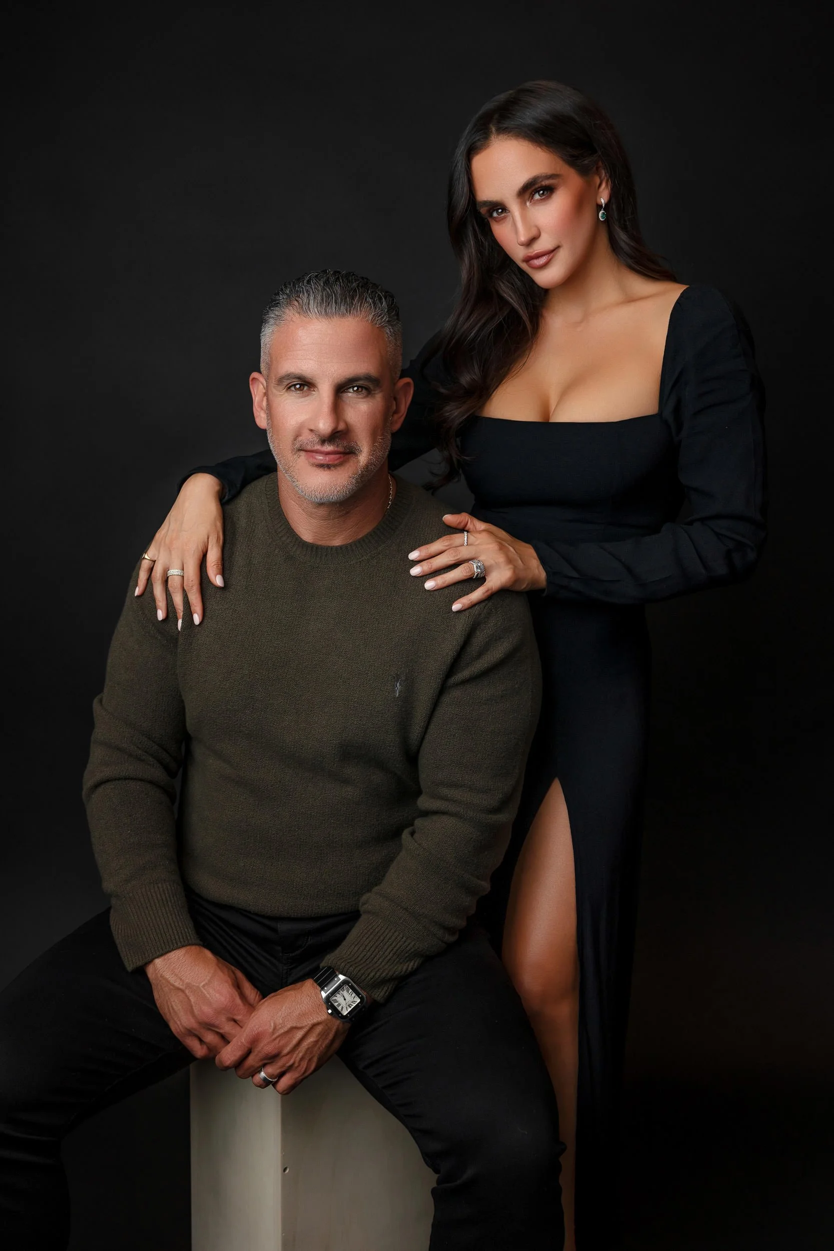 A man with gray hair and a woman with long dark hair posing against a black background. The man is seated, wearing a green sweater and black pants, with rings on his fingers and a watch on his wrist. The woman stands behind him, wearing a black dress