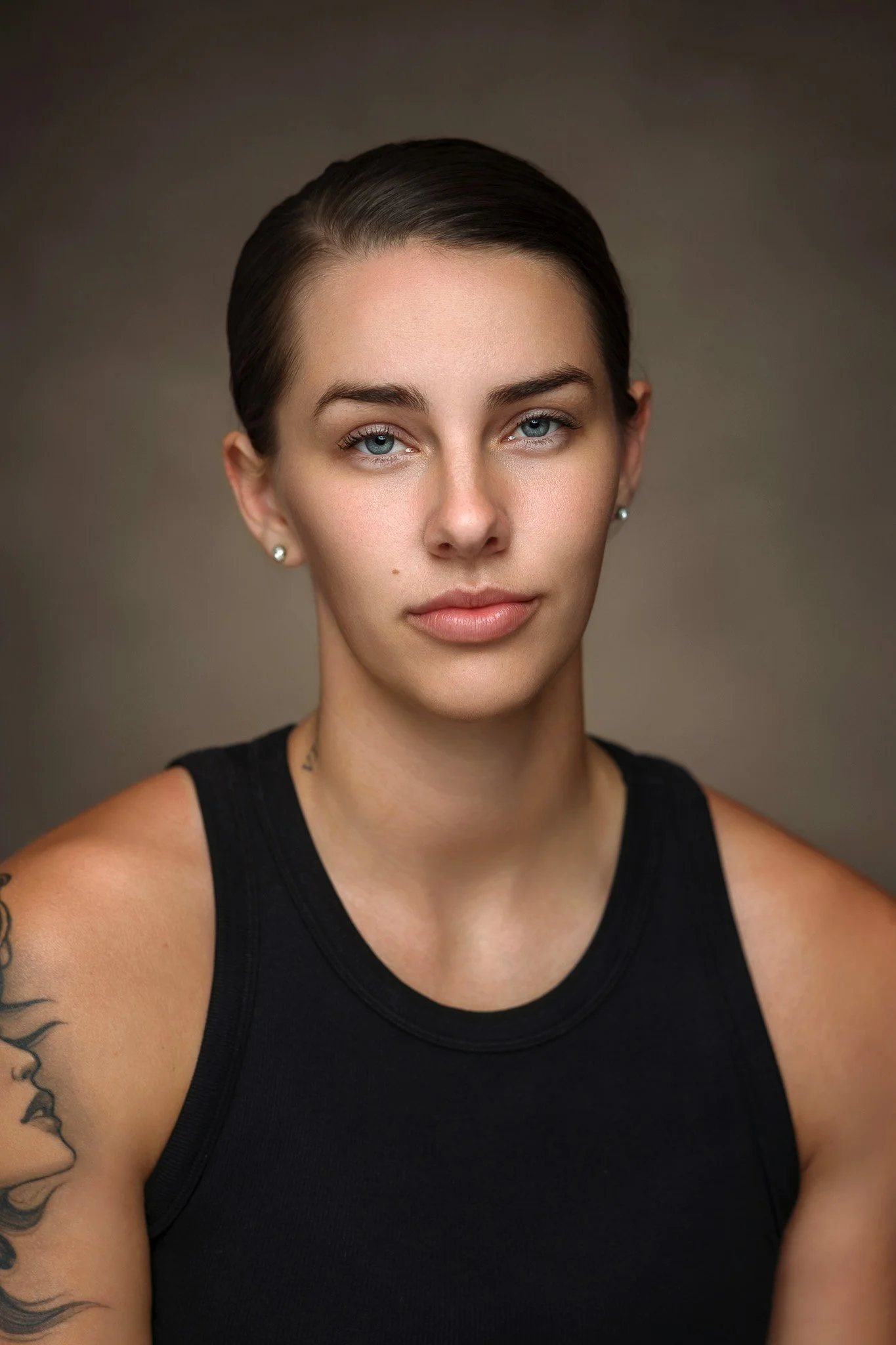 A woman with short dark hair, blue eyes, and light skin wearing a black sleeveless top, earrings, and tattoos on her arm, against a neutral background.