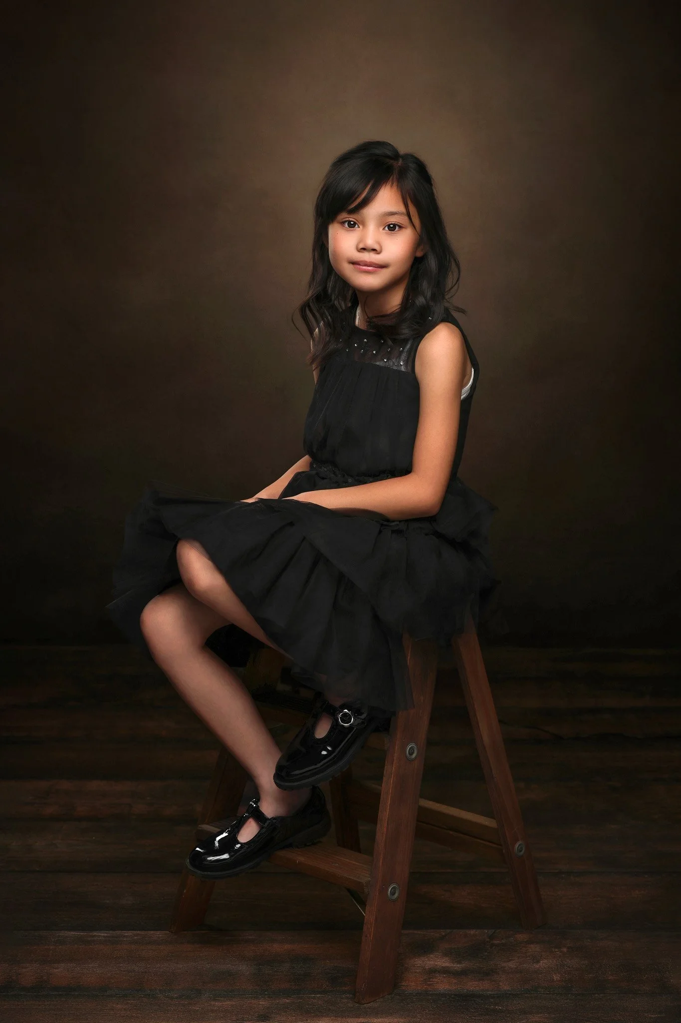 A young girl with shoulder-length black hair, wearing a black dress with a tulle skirt, sitting sideways on a wooden chair with her legs crossed, against a plain dark background.