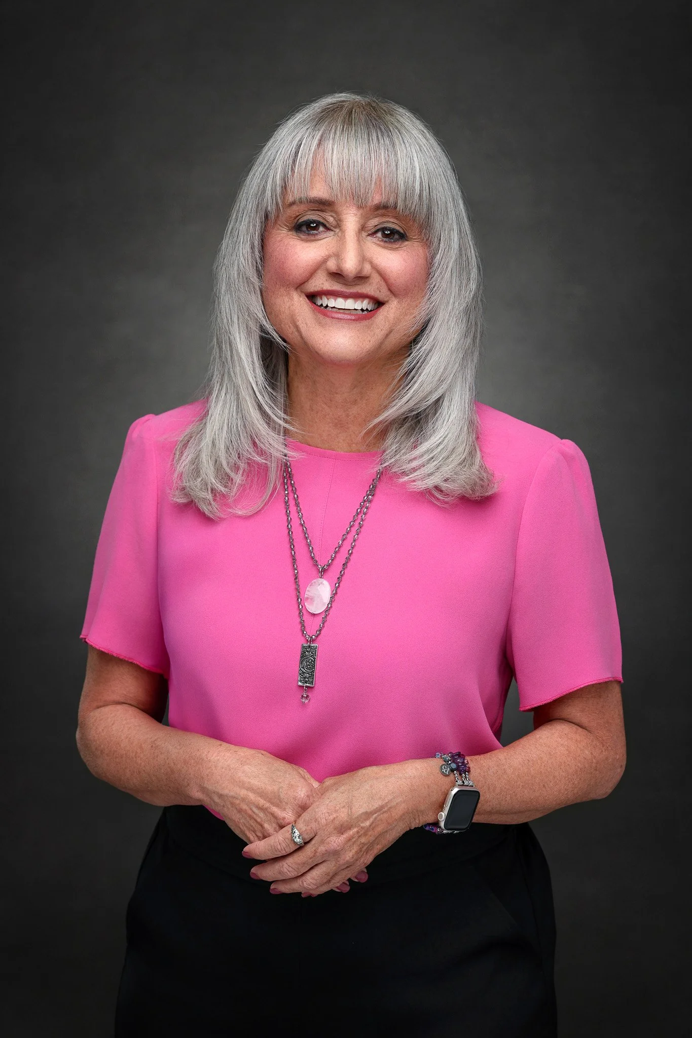 A smiling woman with gray hair in a bob style, wearing a pink top, a silver necklace with a pink stone, a smartwatch, bracelets, a ring, and crossed her hands in front of her against a dark gray background.